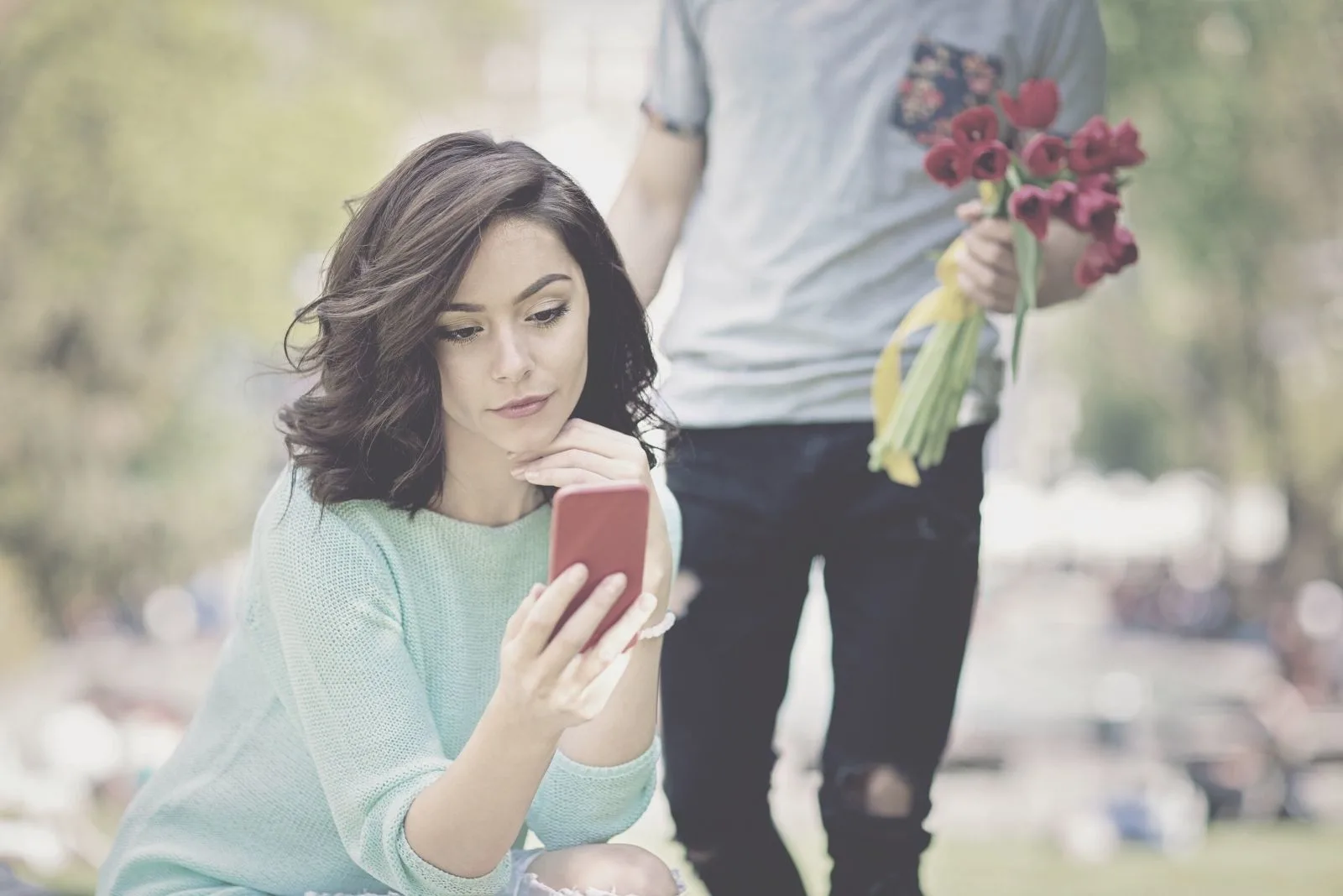 man is bringing flowers to a woman sitting in the park looking at her smartphone