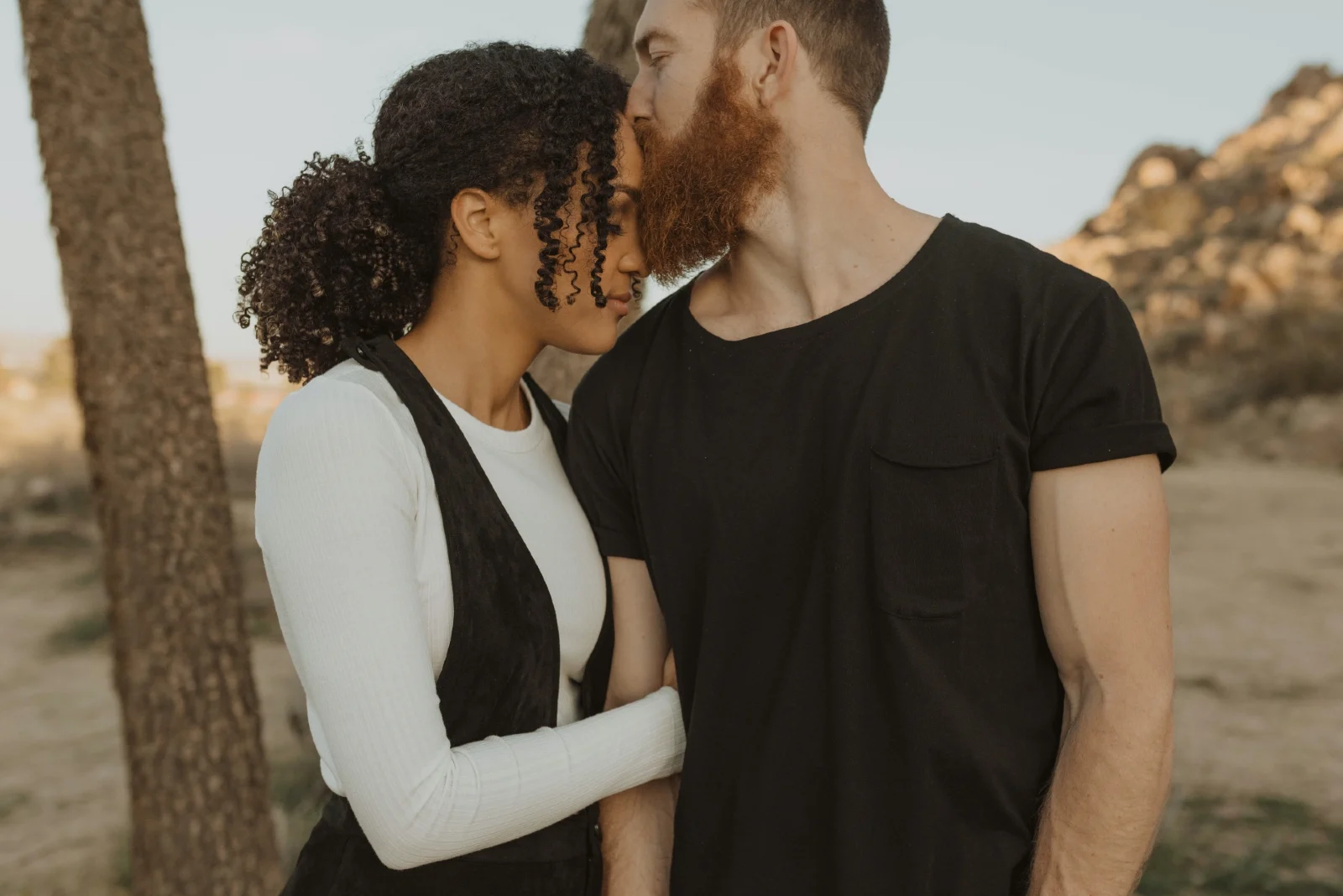 man kissing woman's forehead while standing near tree