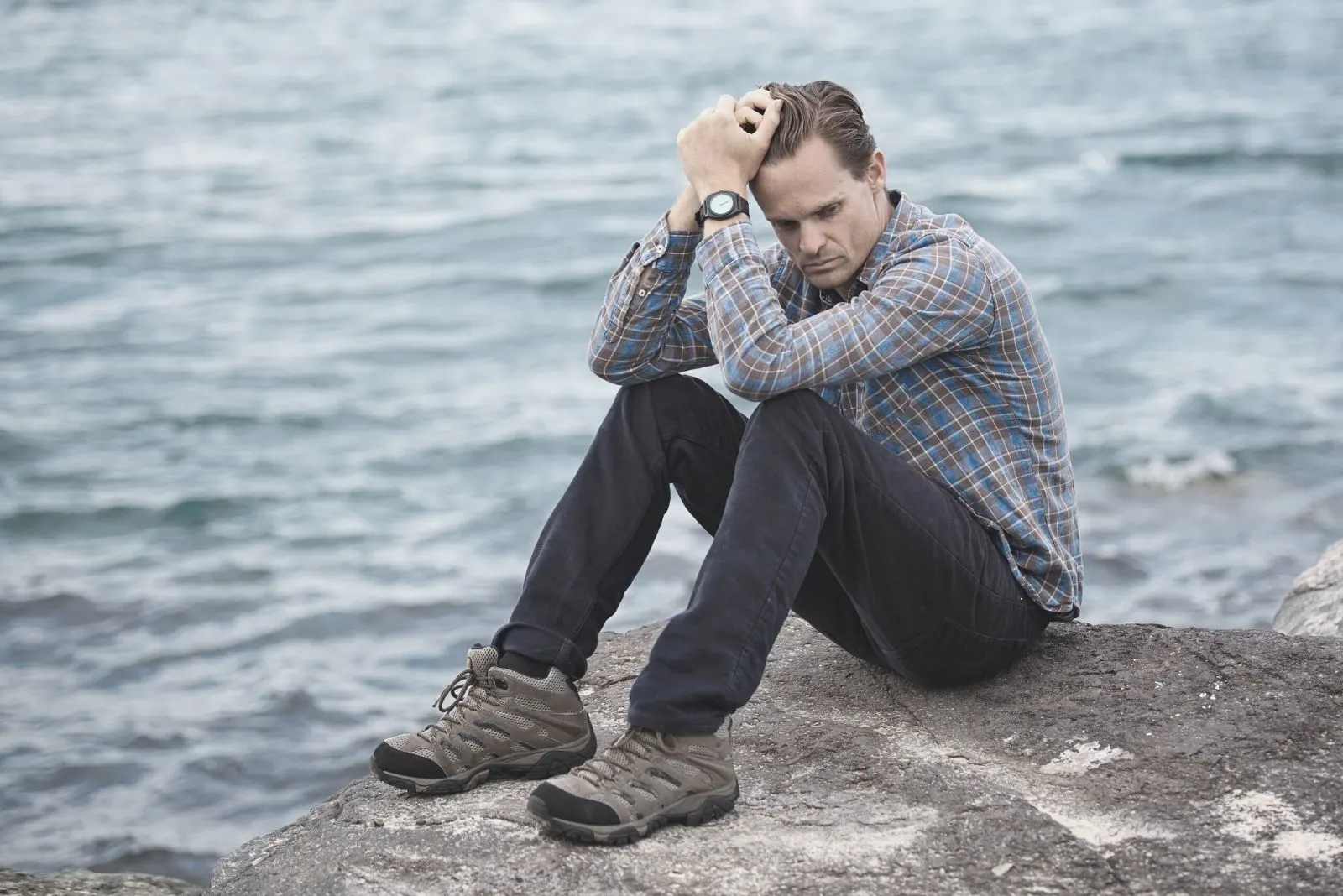 man sitting on the rock near the body of water wearing blue and maroon plaid shirt
