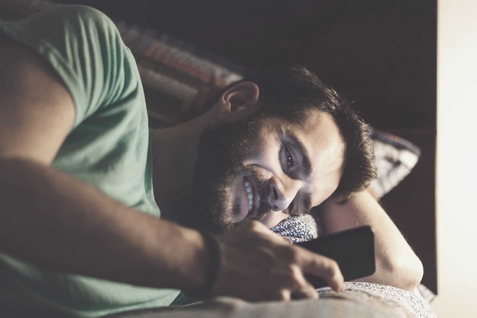 hombre enviando mensajes de texto mientras está acostado en la cama sonriendo y en la habitación oscura