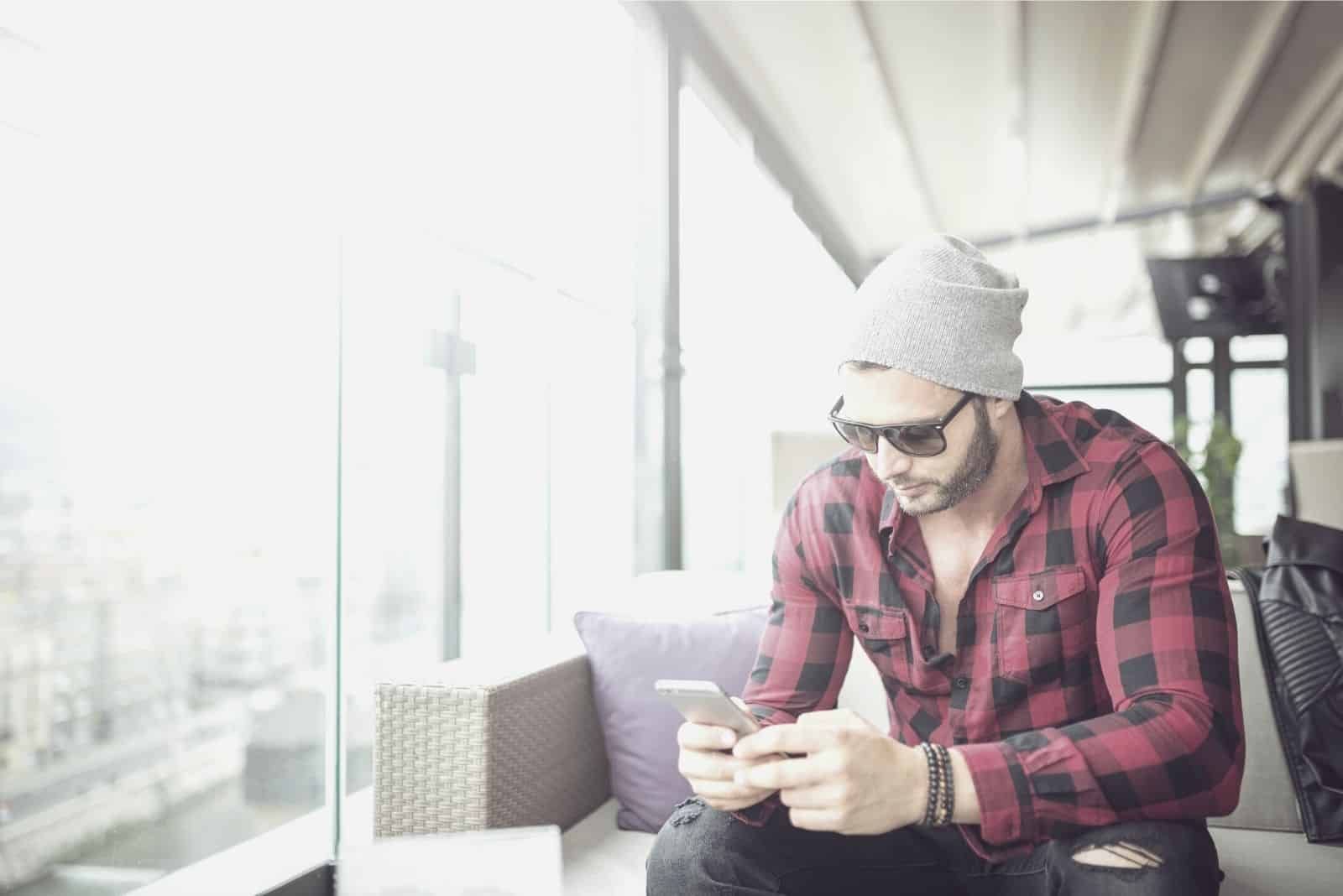 hombre enviando mensajes de texto mientras está sentado dentro de la cafetería con gorro y gafas de sol