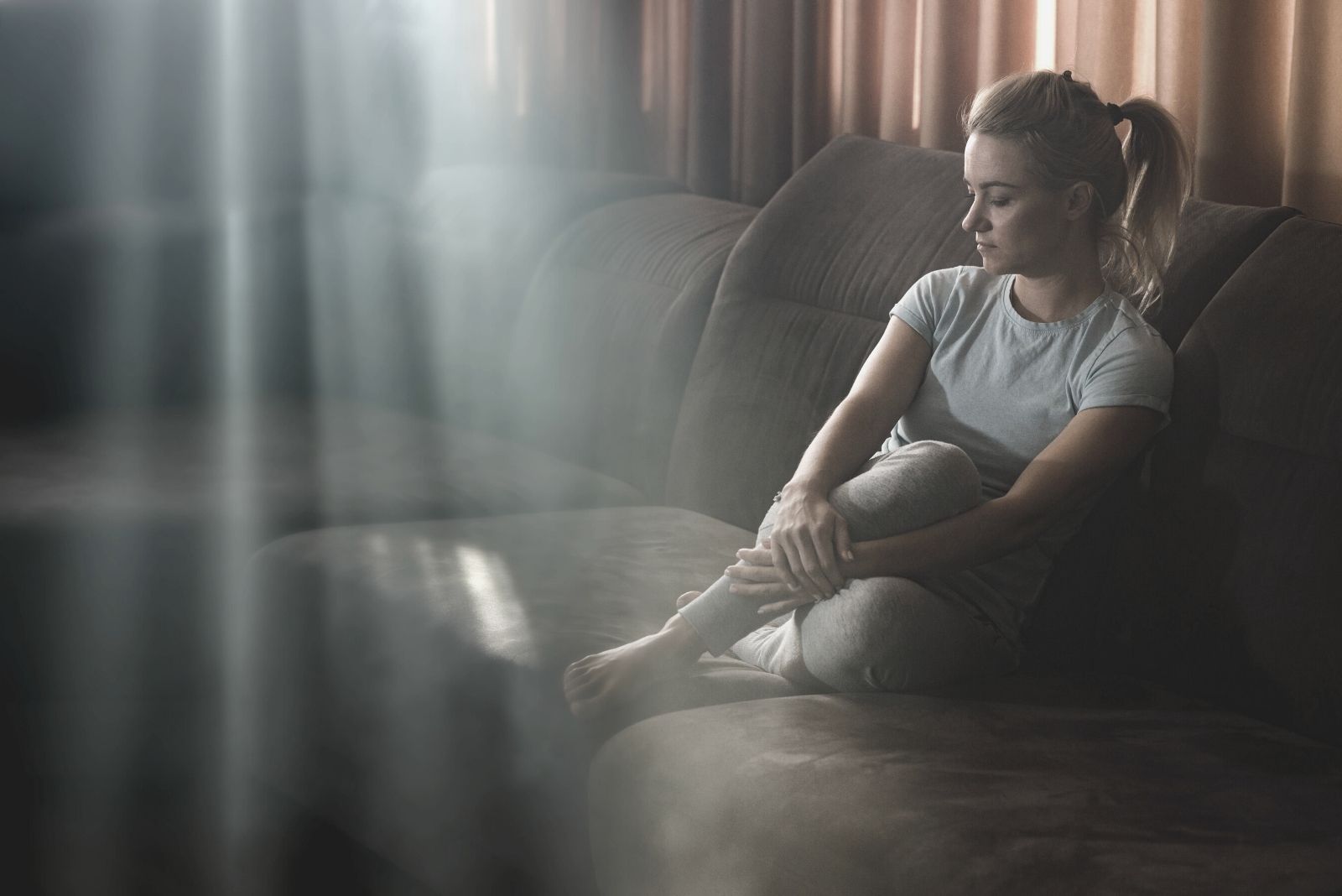 pensive woman feeling sad at home sitting in the dark living room with a few sunrays coming in