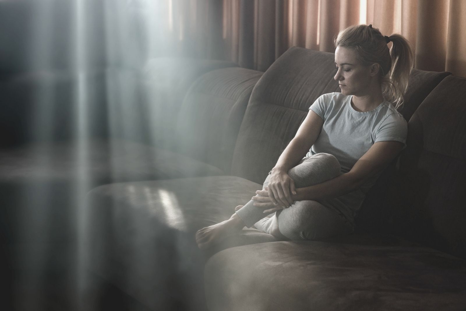 mulher pensativa sentindo-se triste em casa, sentada na sala de estar escura com alguns raios de sol a entrar