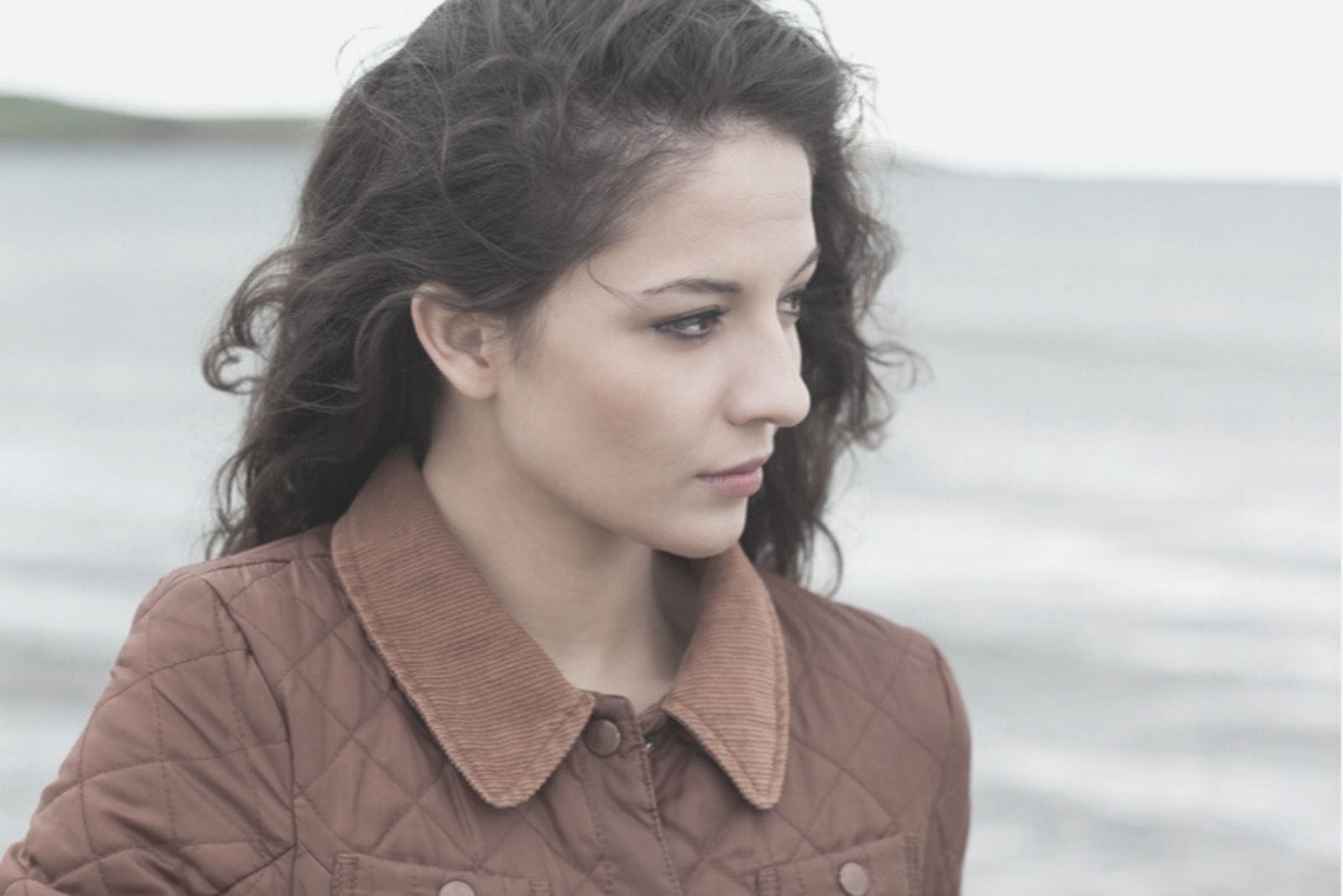 Donna pensierosa che cammina in riva al mare indossando una giacca marrone in una fotografia ravvicinata