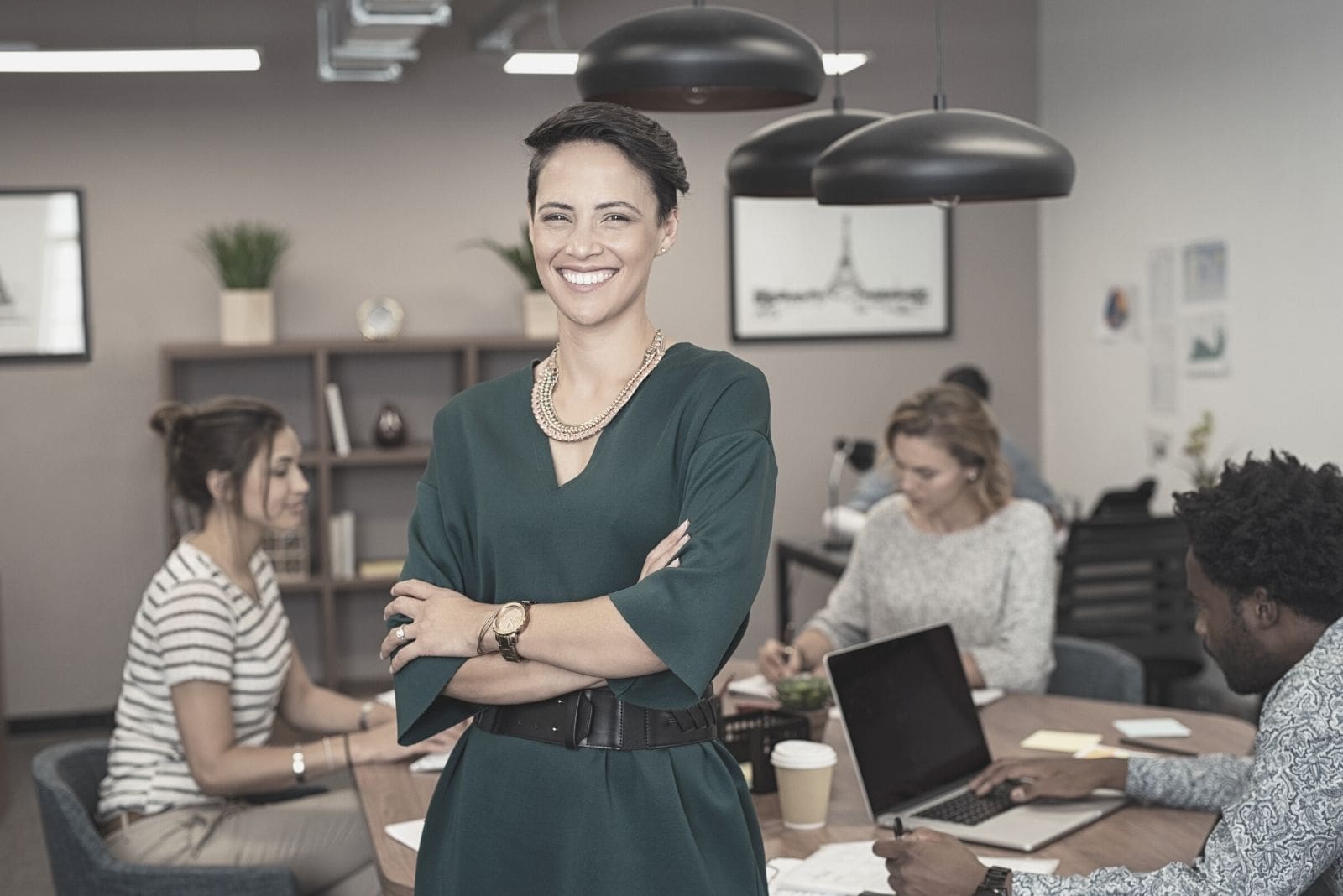 retrato de uma mulher bem sucedida, de pé, com colegas a trabalhar ao fundo, no interior de um escritório