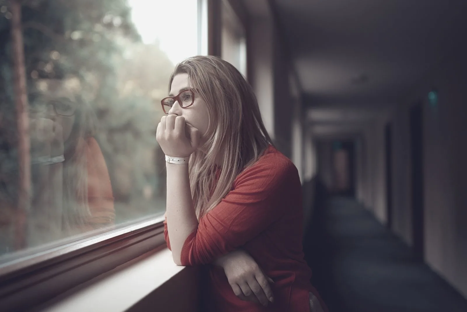 side view of a pensive woman looking outside the window wearing eyeglasses and leaning on the window sill