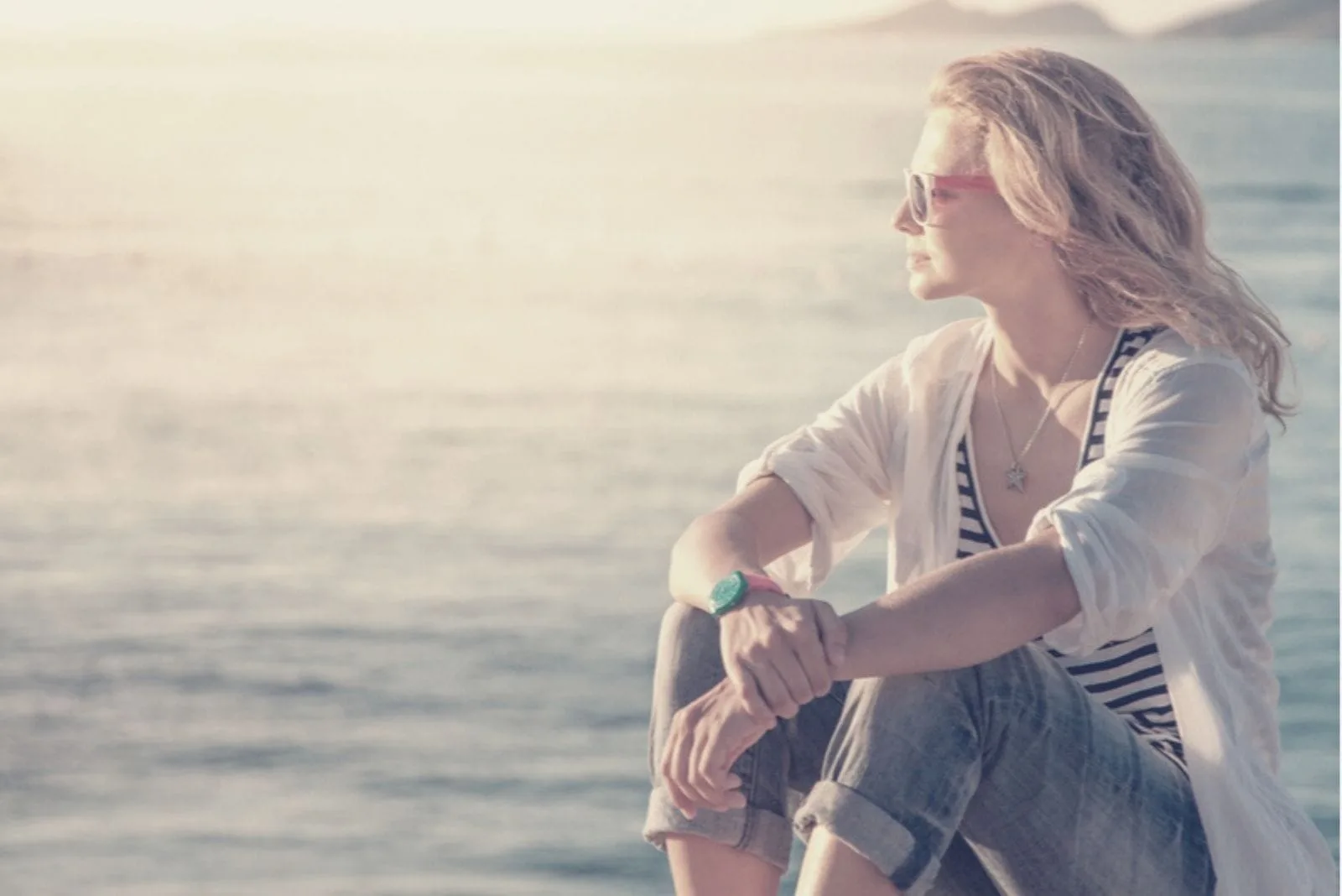 thoughtful woman sitting near the body of water wearing shades and facing the setting sun