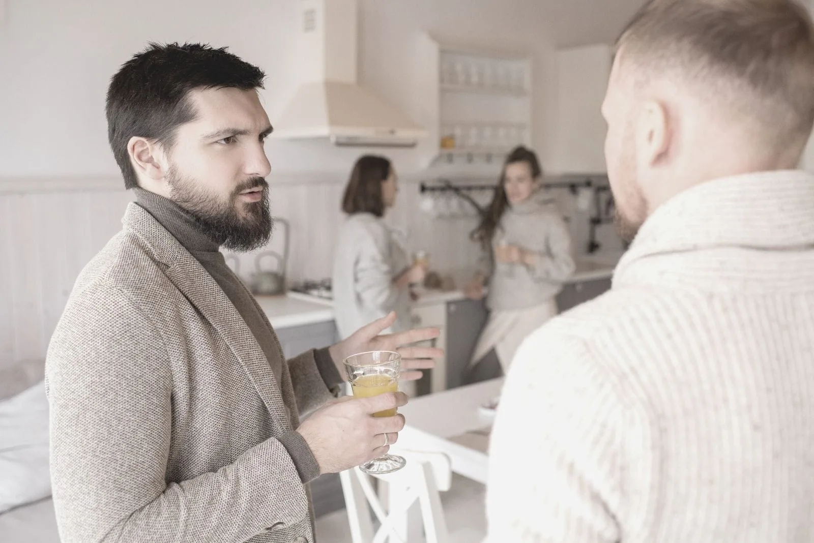 two men talking seriously inside the kitchen with two women talking at the background