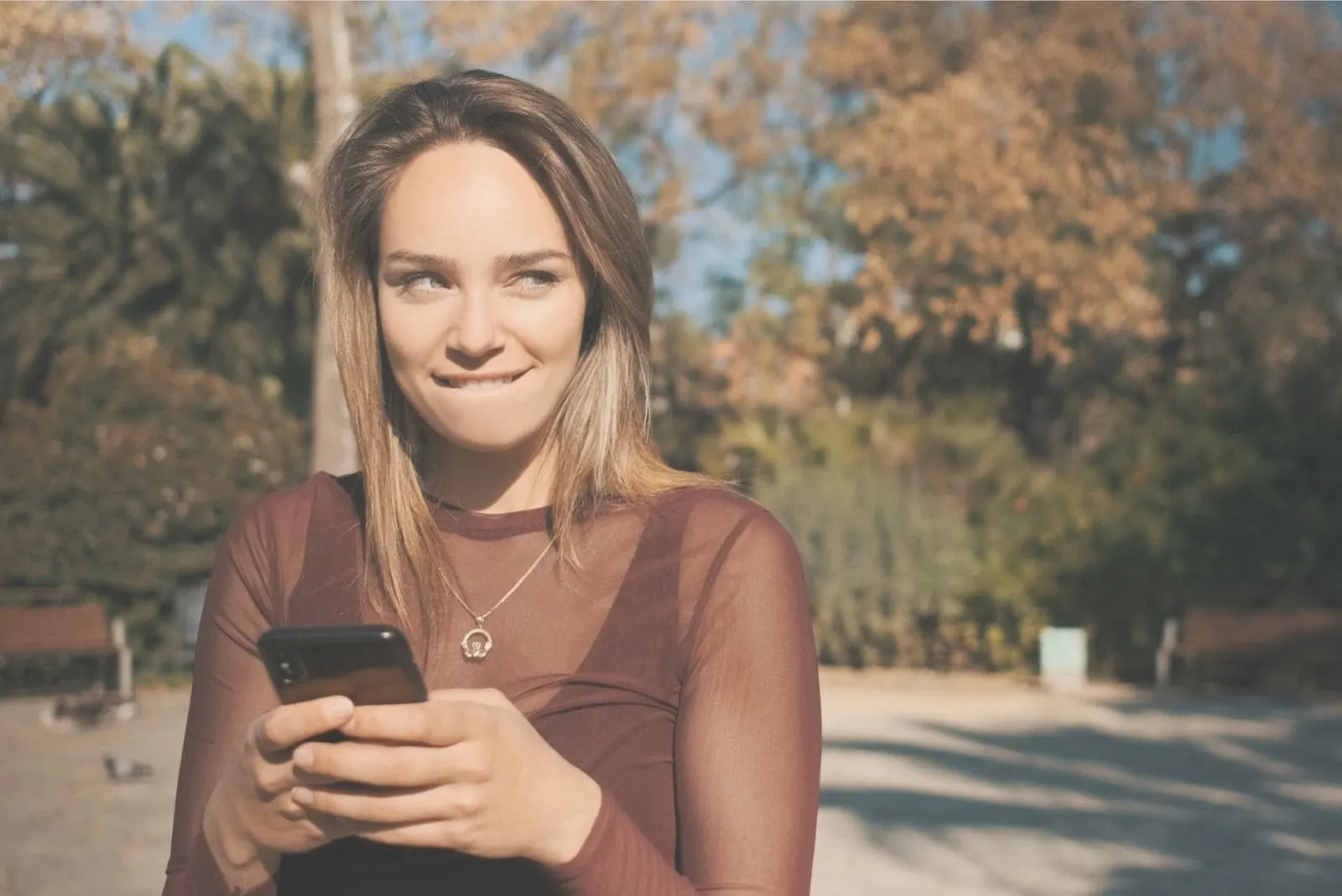 woman biting lip while texting and looking away standing outdoors