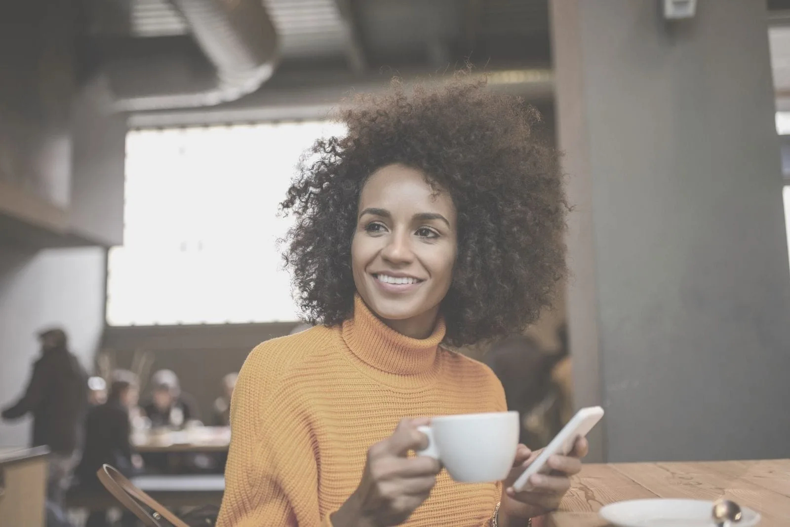 woman drinking coffee inside a cafe and holding a cellphone looking away and smiling