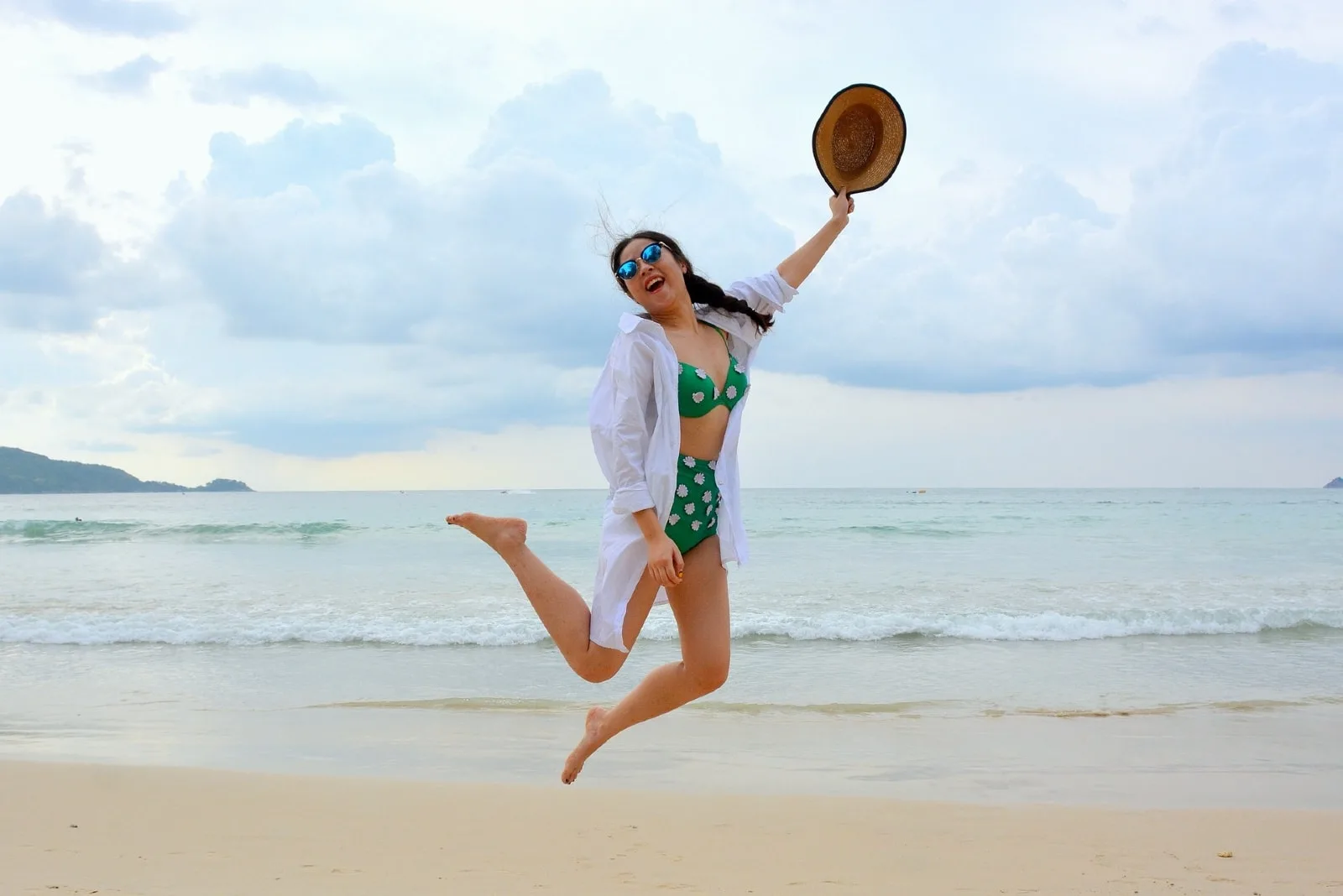 woman holding hat while jumping on seashore