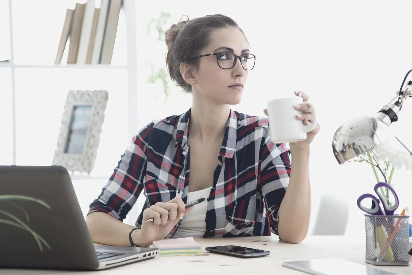 woman sitting in office thinking and looking away wearing eyeglasses and drinking from a mug