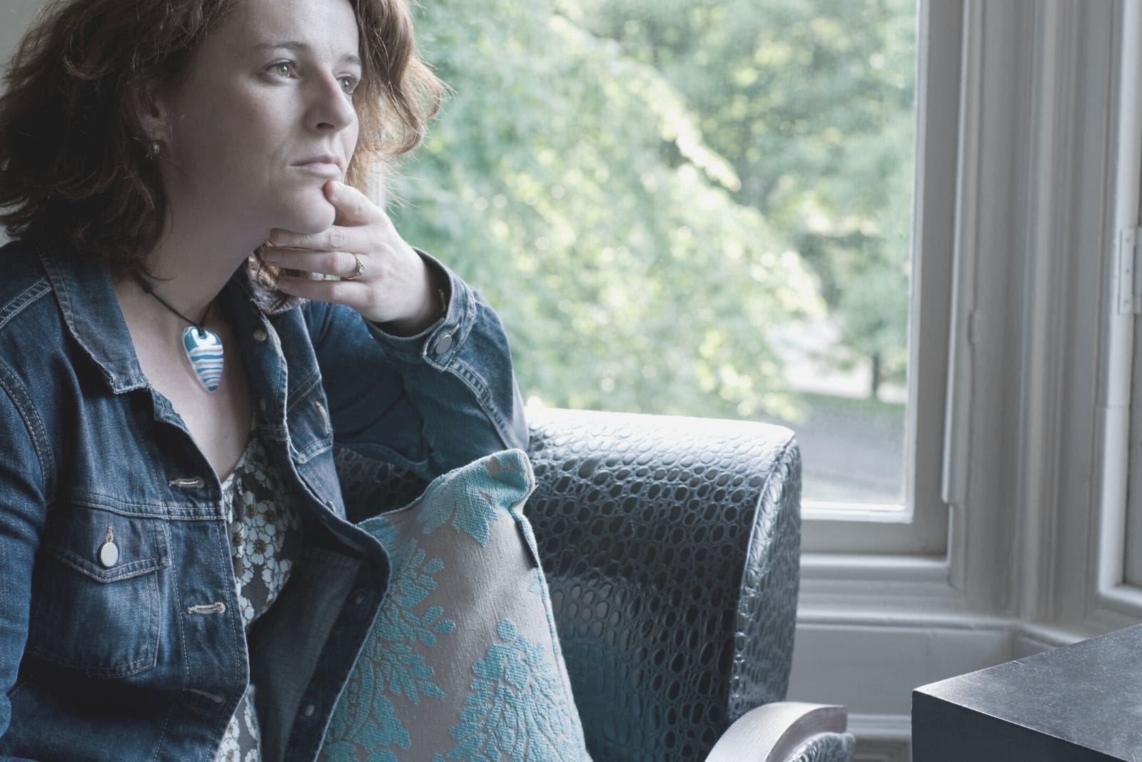donna con giacca di jeans pensierosa e seduta vicino alle finestre
