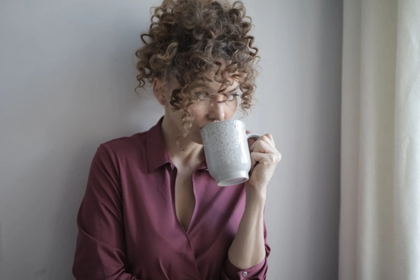 woman with curly hair leaning on the room's wall looking away and drinking from a mug
