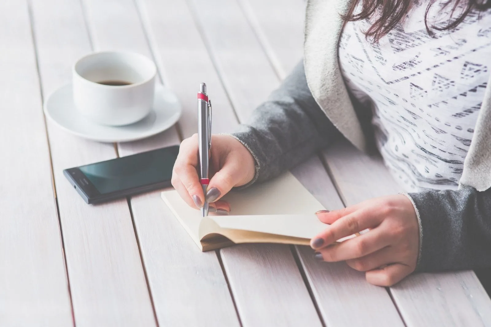 woman writing on her journal on a wooden table with phone and coffee on the table in cropped image
