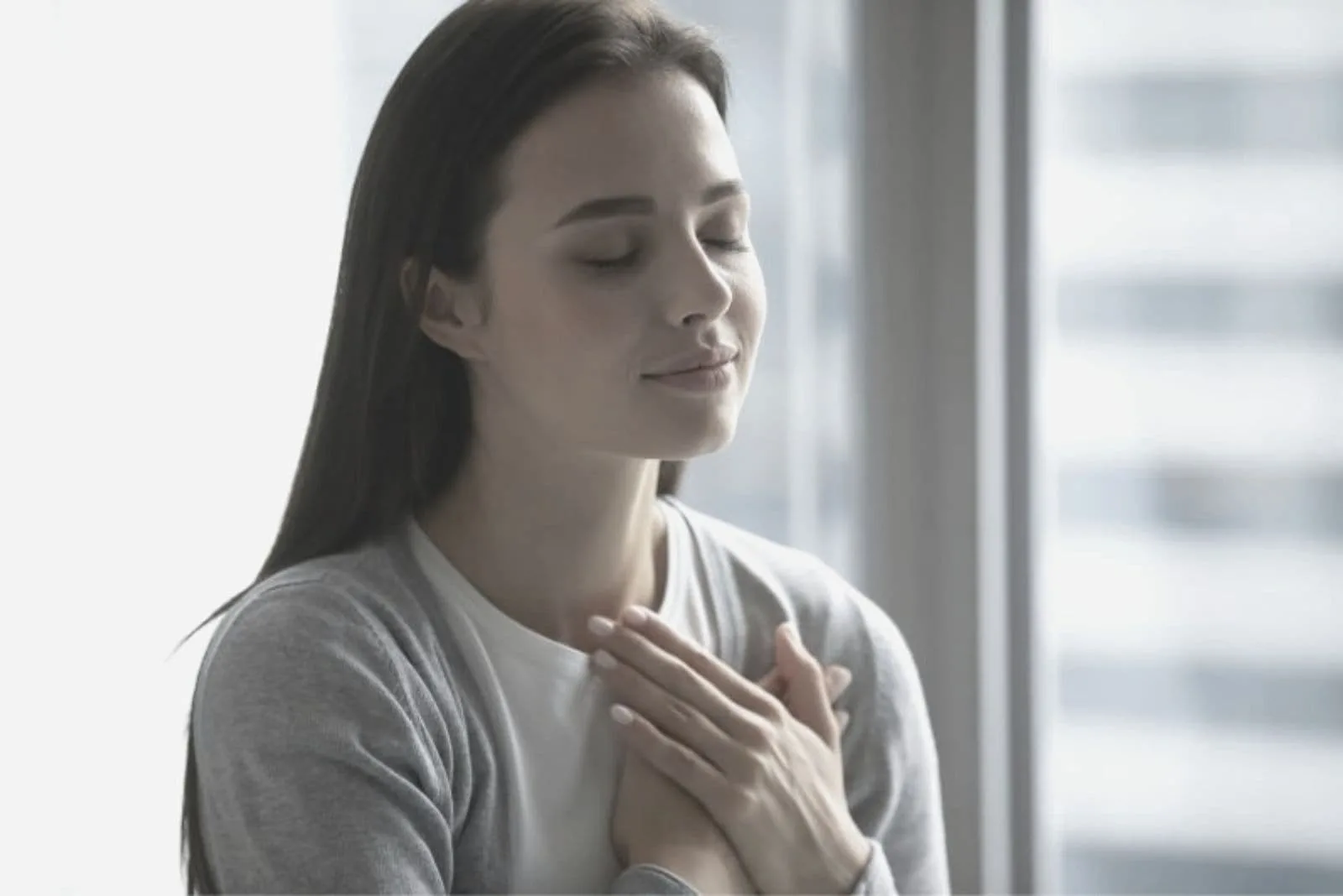 young beautiful woman closing her eyes and putting her hands close to her chest standing indoors