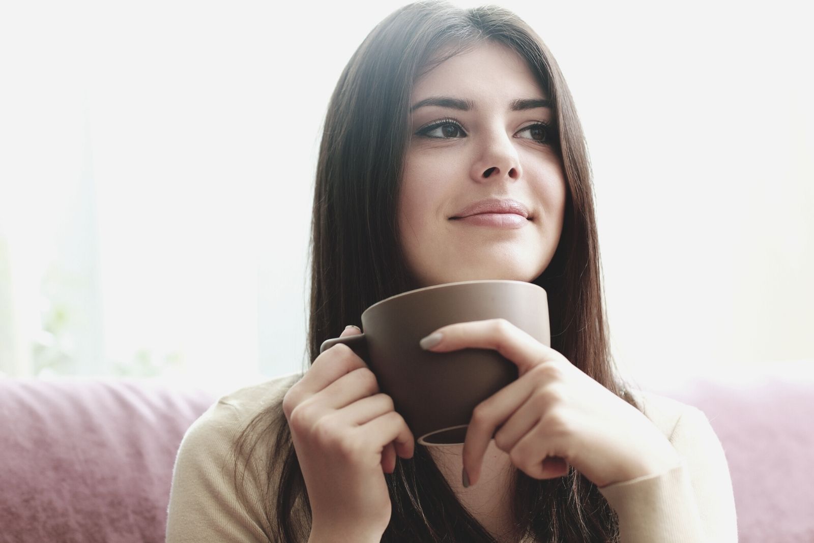 giovane donna felice e riflessiva che beve un caffè e si siede sul divano in una fotografia ravvicinata