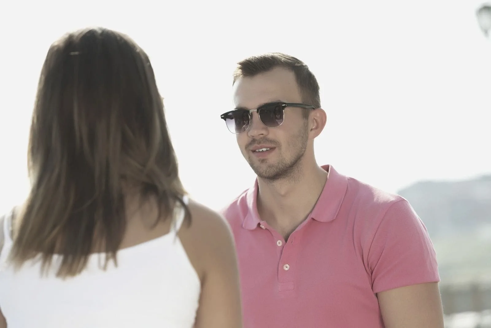 young man wearing pink polo shirt and sunglasses while facing and talking to a woman outdoors