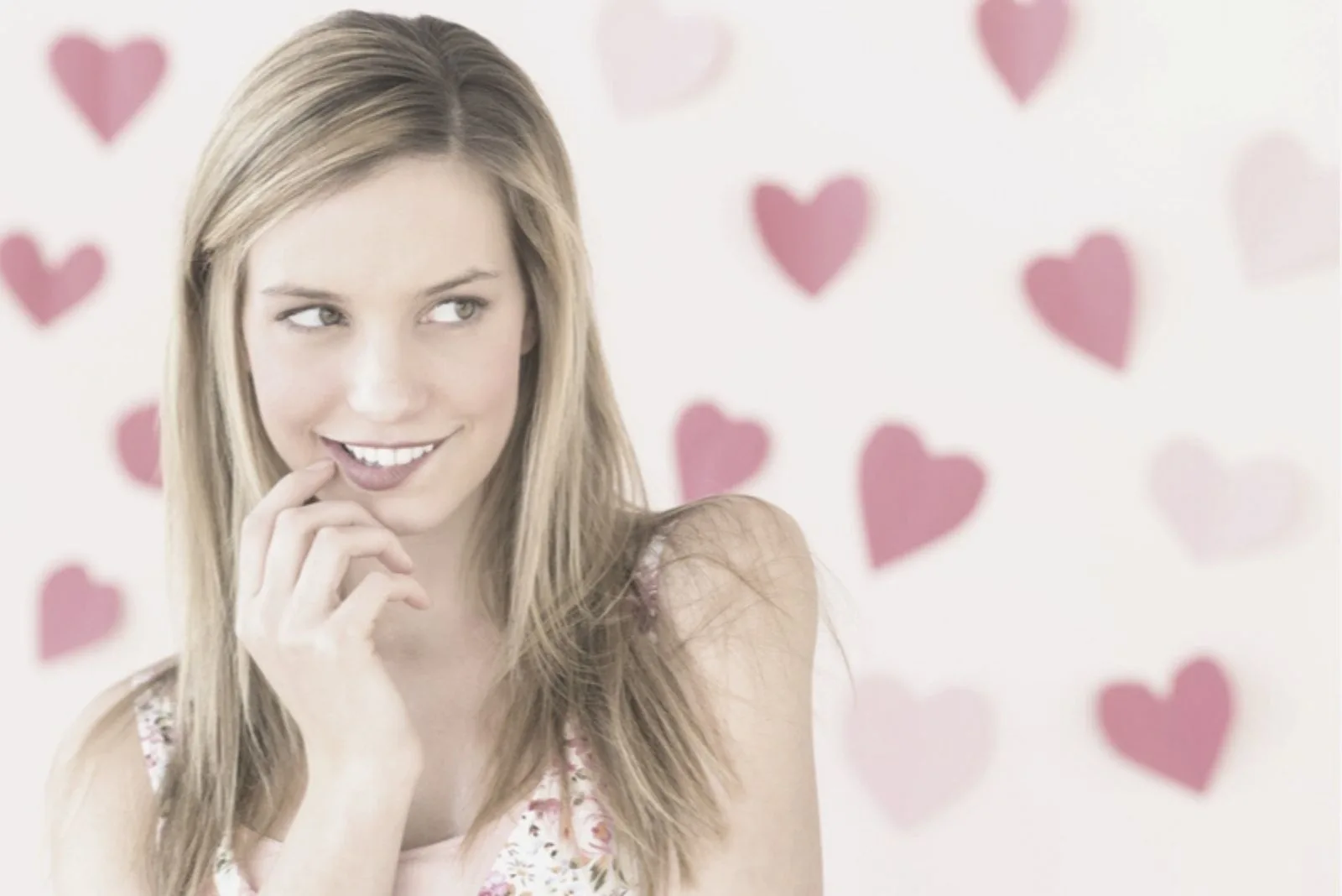 young woman biting lip and looking away standing near the white wall with pink hearts