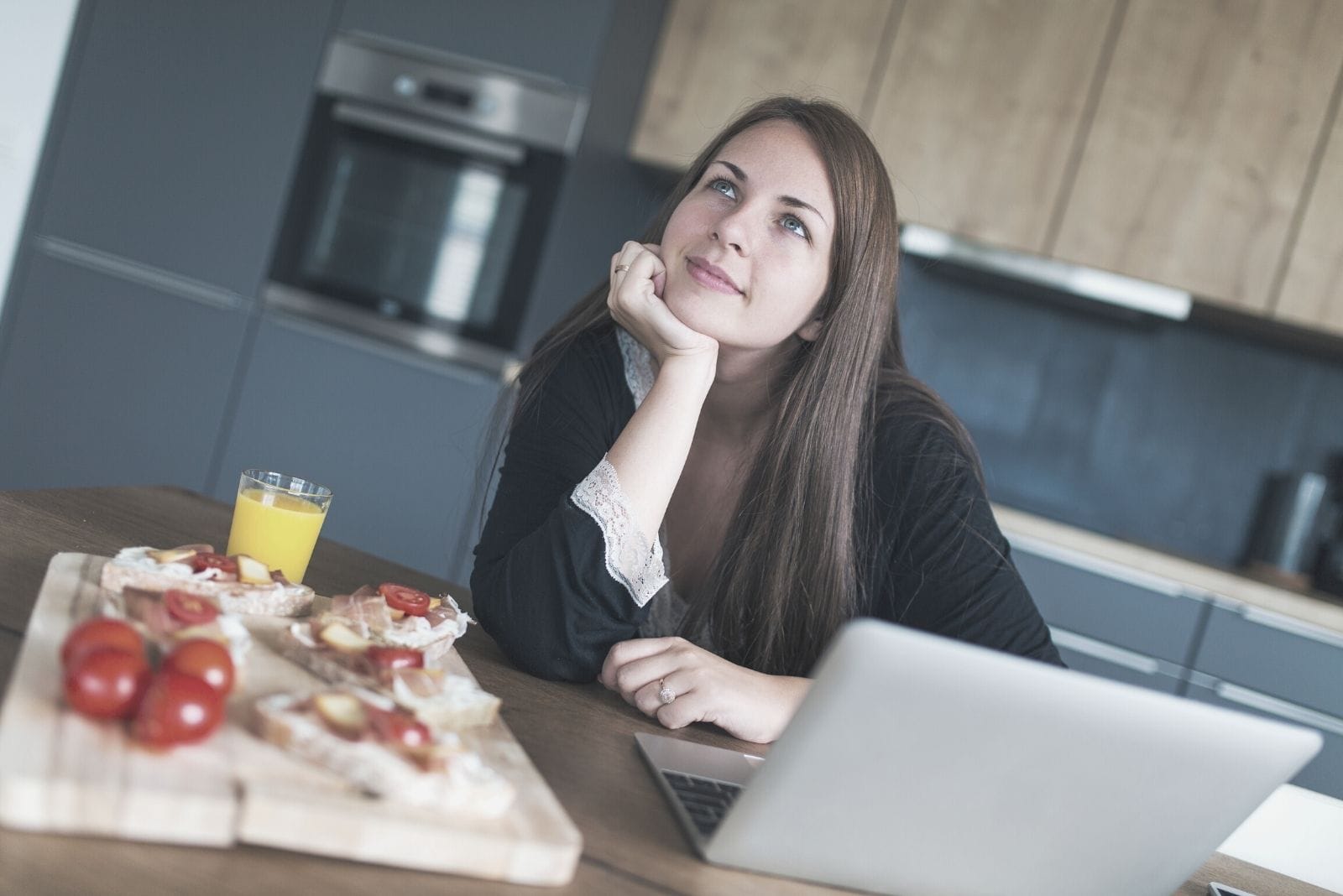 young woman daydreaming in the kitchen with food on the side while working on laptop