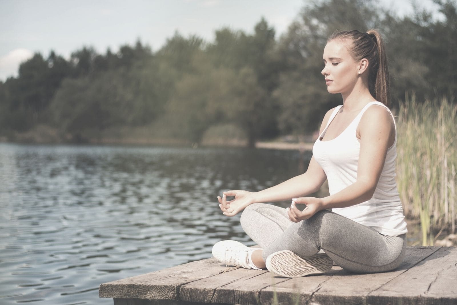 giovane donna che medita in posizione yoga in cima a una piattaforma di legno vicino a uno specchio d'acqua