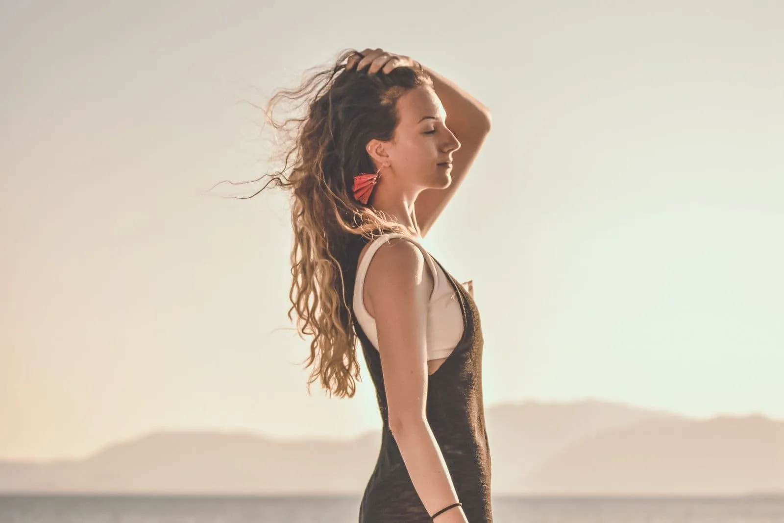 young woman posing while touching her hair and closing her eyes standing on the shore