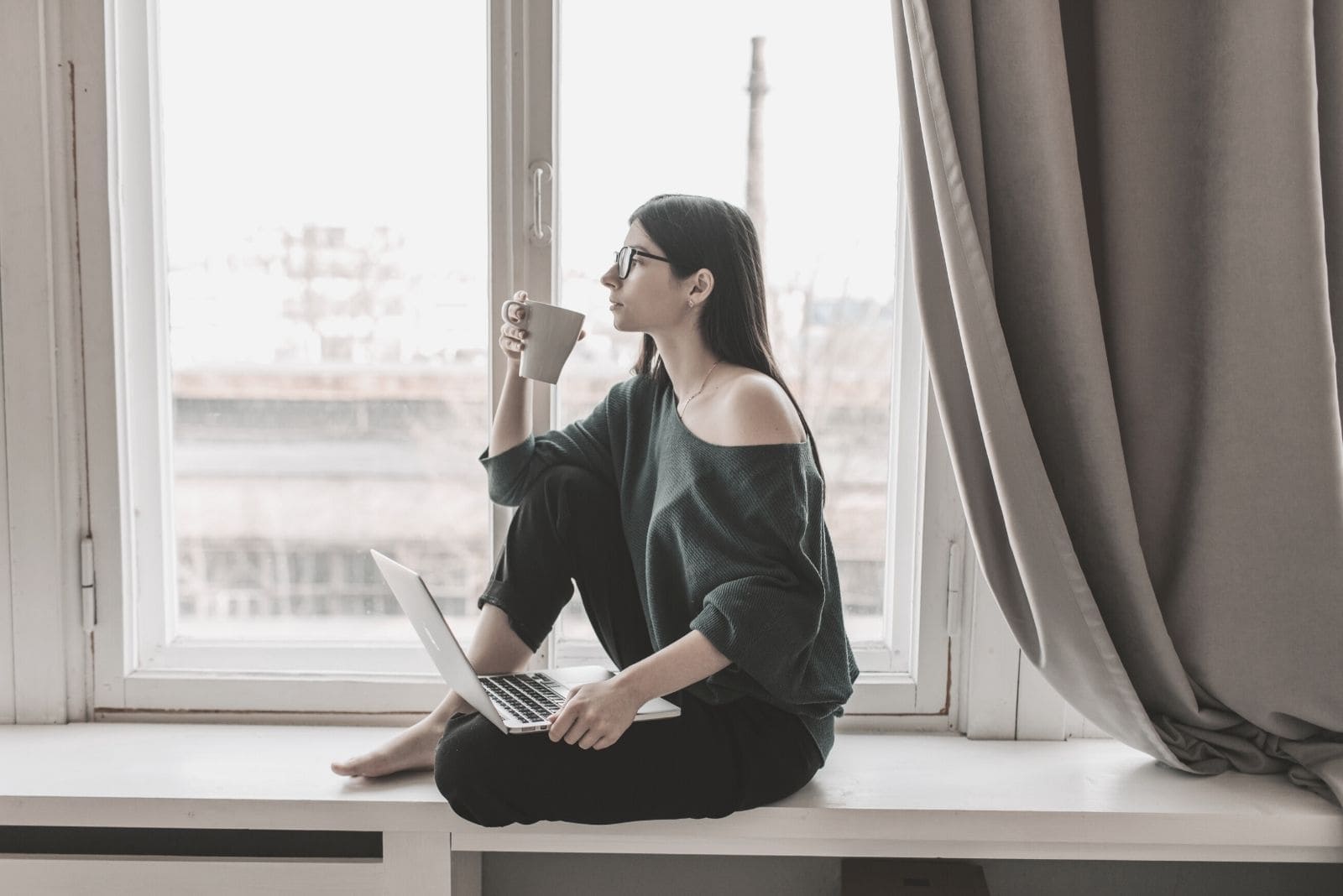young woman thinking drinking coffee sitting by the window sill with a laptop on her lap