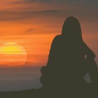 vista posteriore di una donna che guarda il tramonto da sola, seduta vicino allo specchio d'acqua