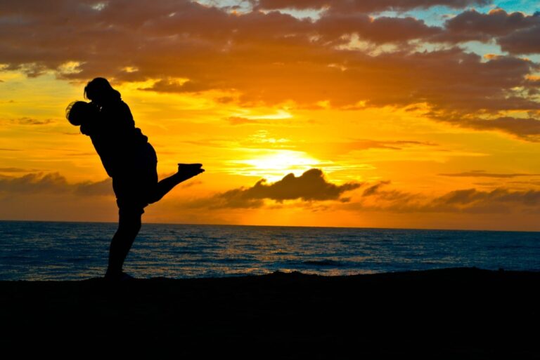 man and woman kissing while standing on shore during sunset