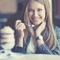 mulher gira a sorrir enquanto come um gelado e olha para alguém dentro do café e com uma mão a prender o cabelo