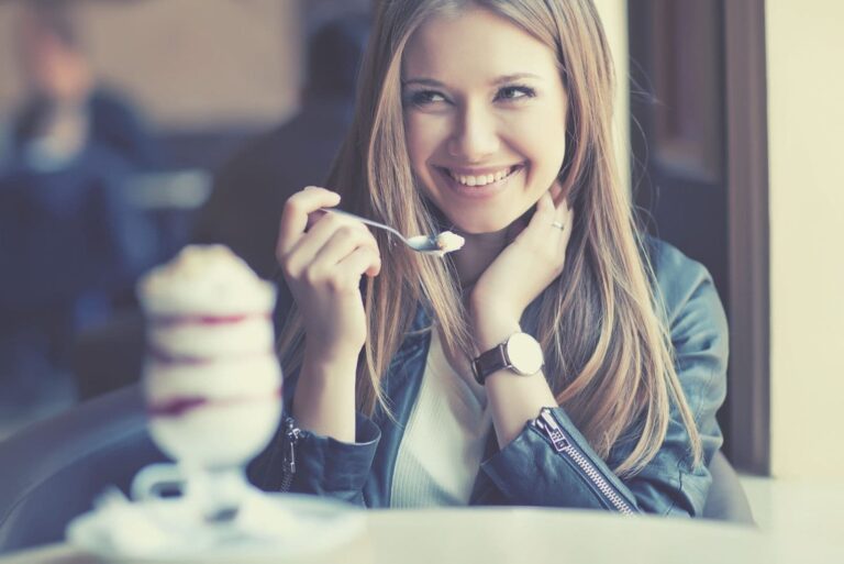 cute woman smiling while eating ice cream and looking at someone inside the cafe and one hand keeping her hair