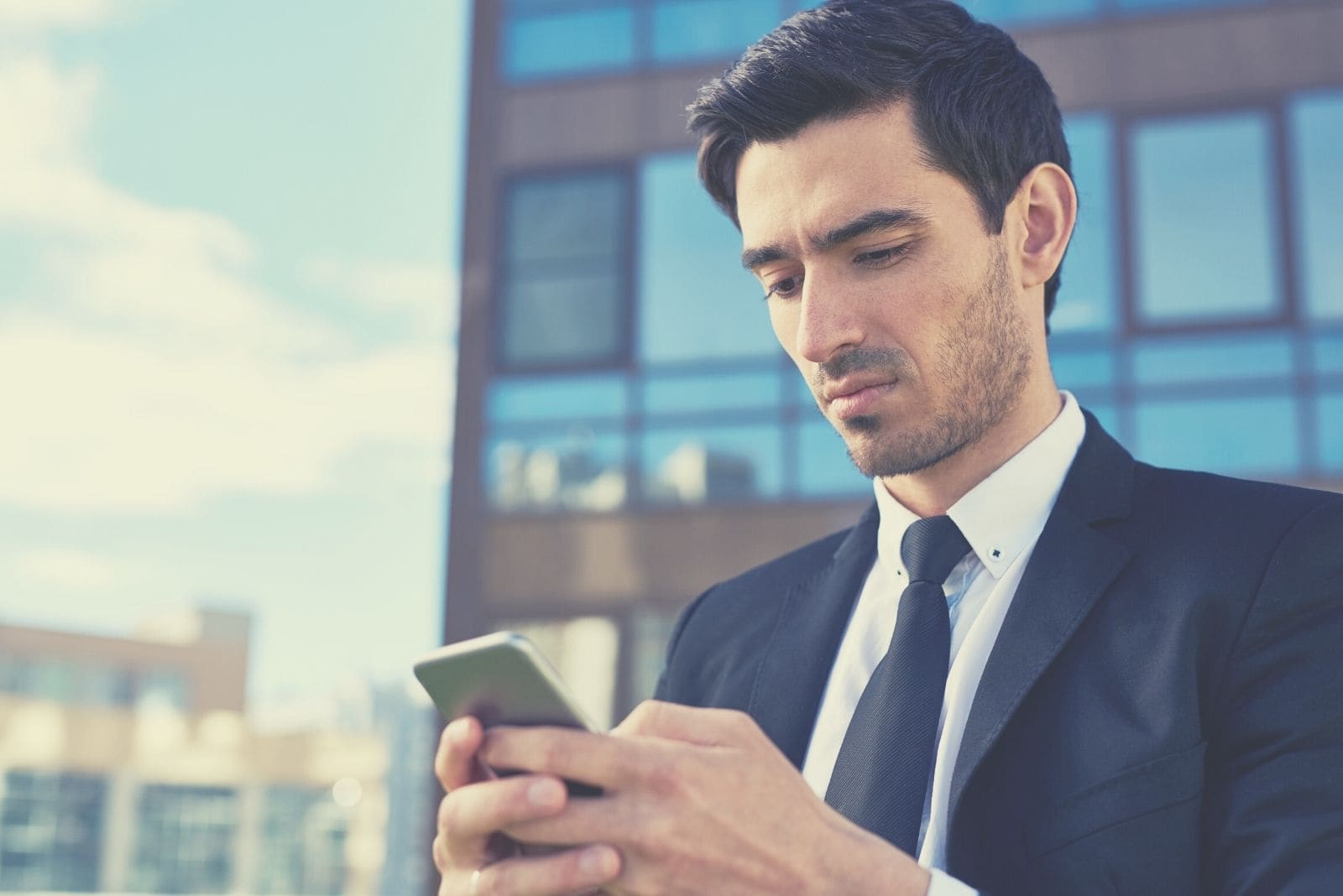 uomo infastidito che risponde a un messaggio di testo dal suo smartphone indossando abiti da lavoro in piedi fuori dall'edificio dell'ufficio