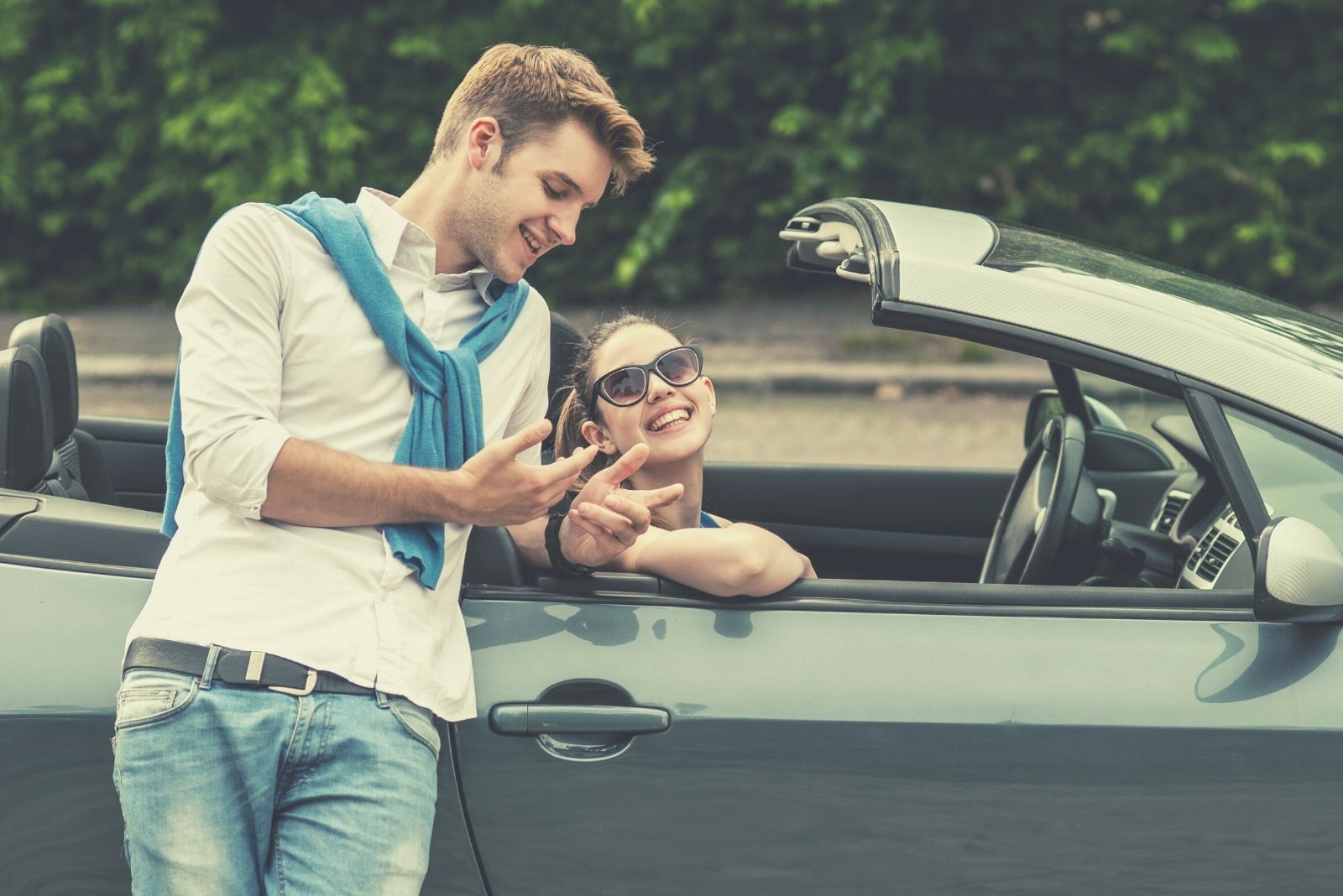 cheerful couple talking outdoors with woman sitting on the car and the man leaning on the topdown's door