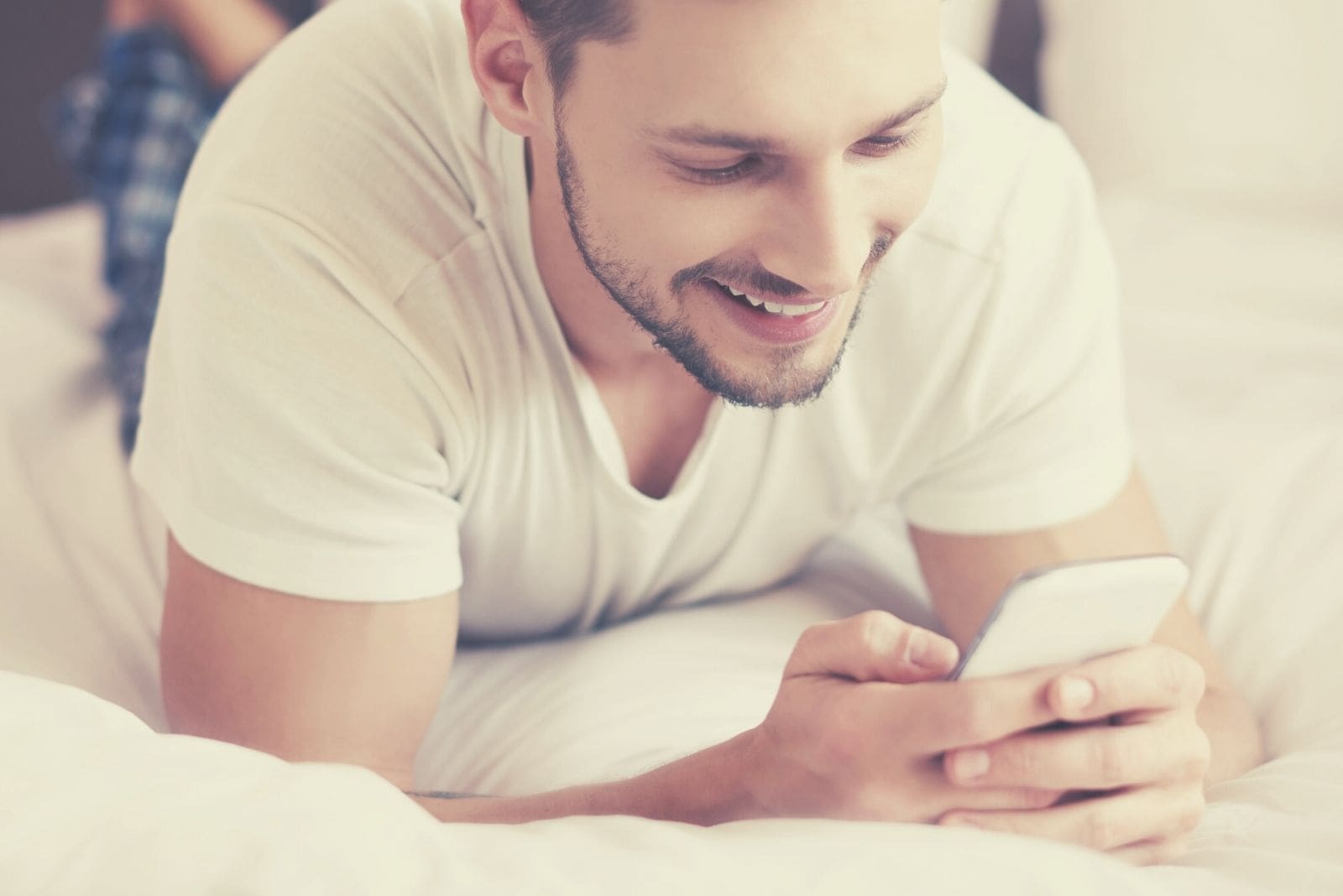 cheerful man texting something funny while lying in the bed in his pajamas