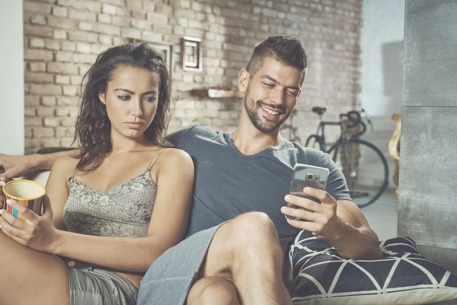 jealous woman sitting beside a happy man texting with legs crossed inside the living room