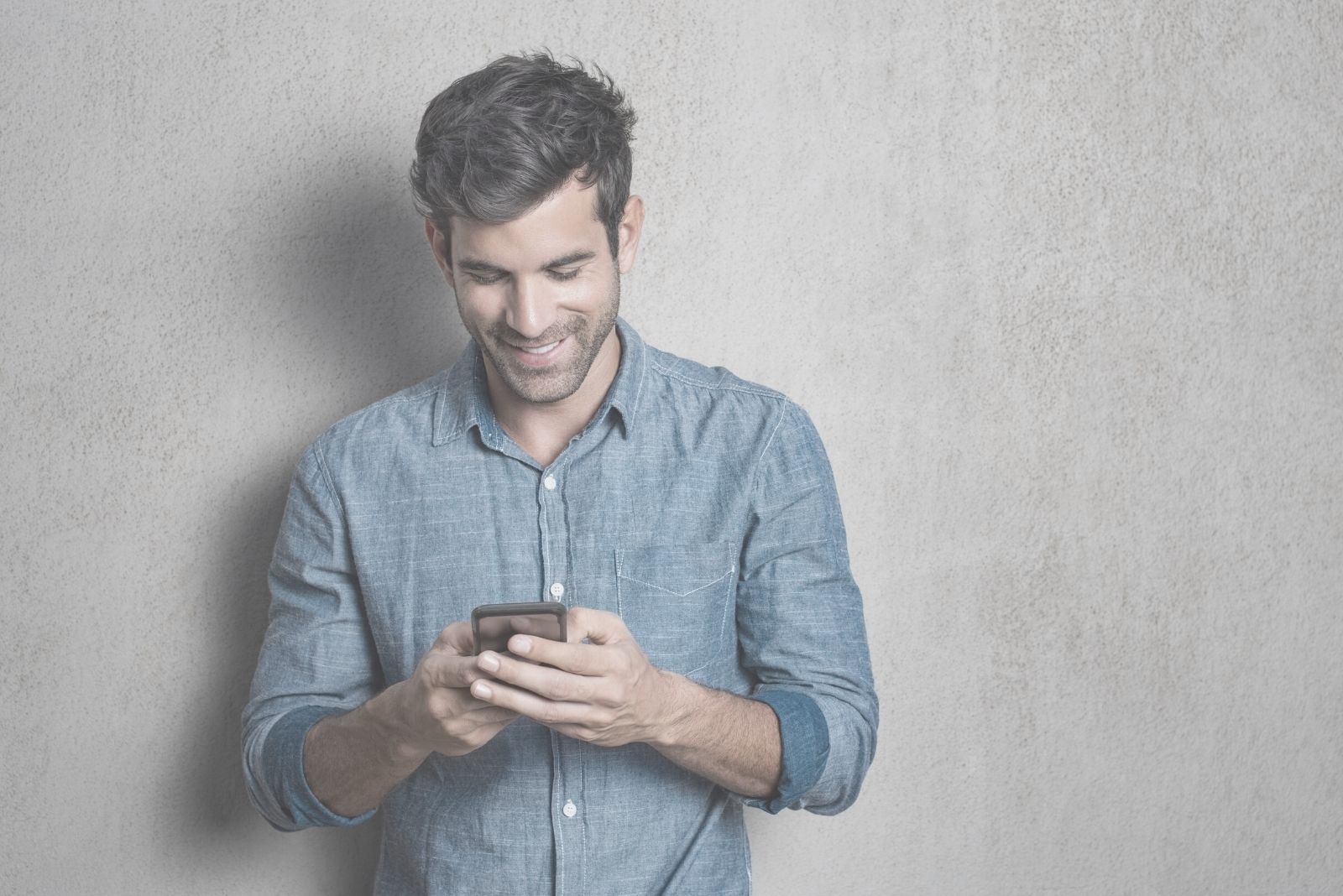 hombre enviando mensajes de texto y sonriendo y de pie apoyado en la pared blanca