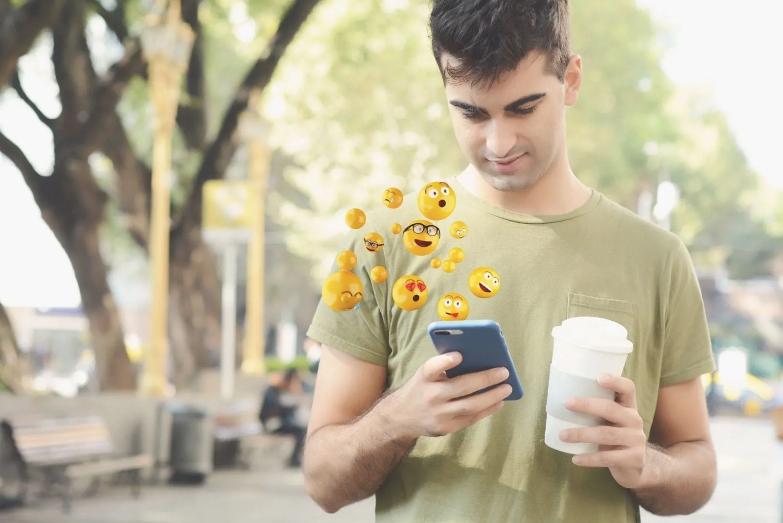 man using smartphone sending emojis and holding cup of coffee while walking in the park