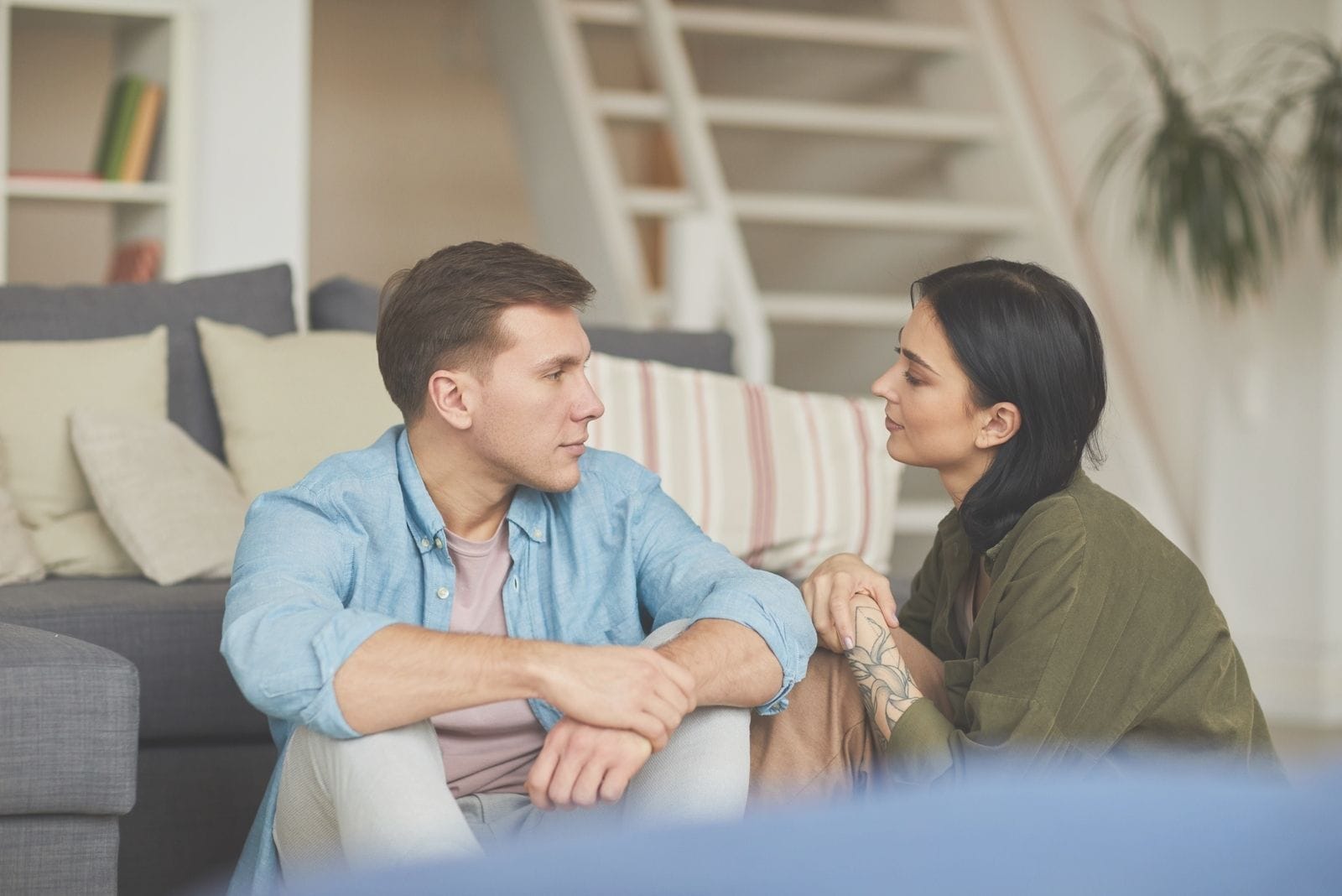 joven pareja moderna hablando el uno al otro sinceramente mientras se está sentado cómodamente en el suelo cerca del sofá dentro de la sala de estar