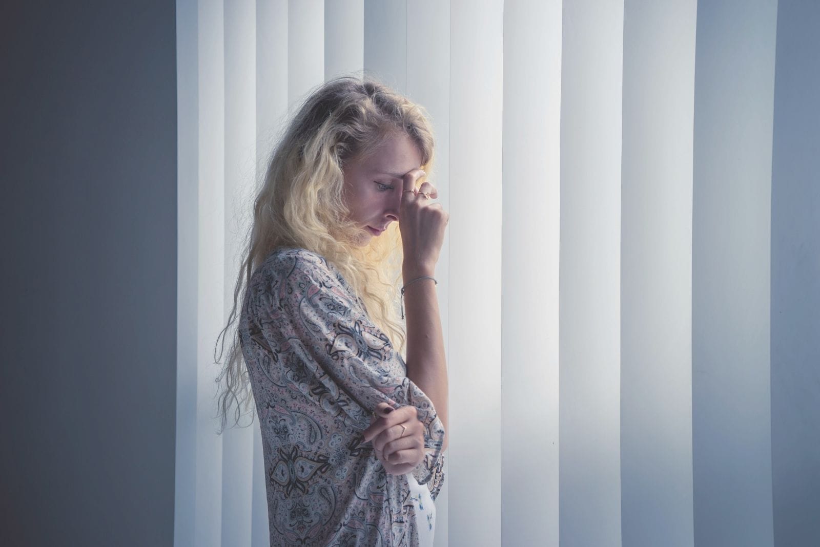 pensive blonde caucasian woman placing her hand in her forehead standing near the curtained windows