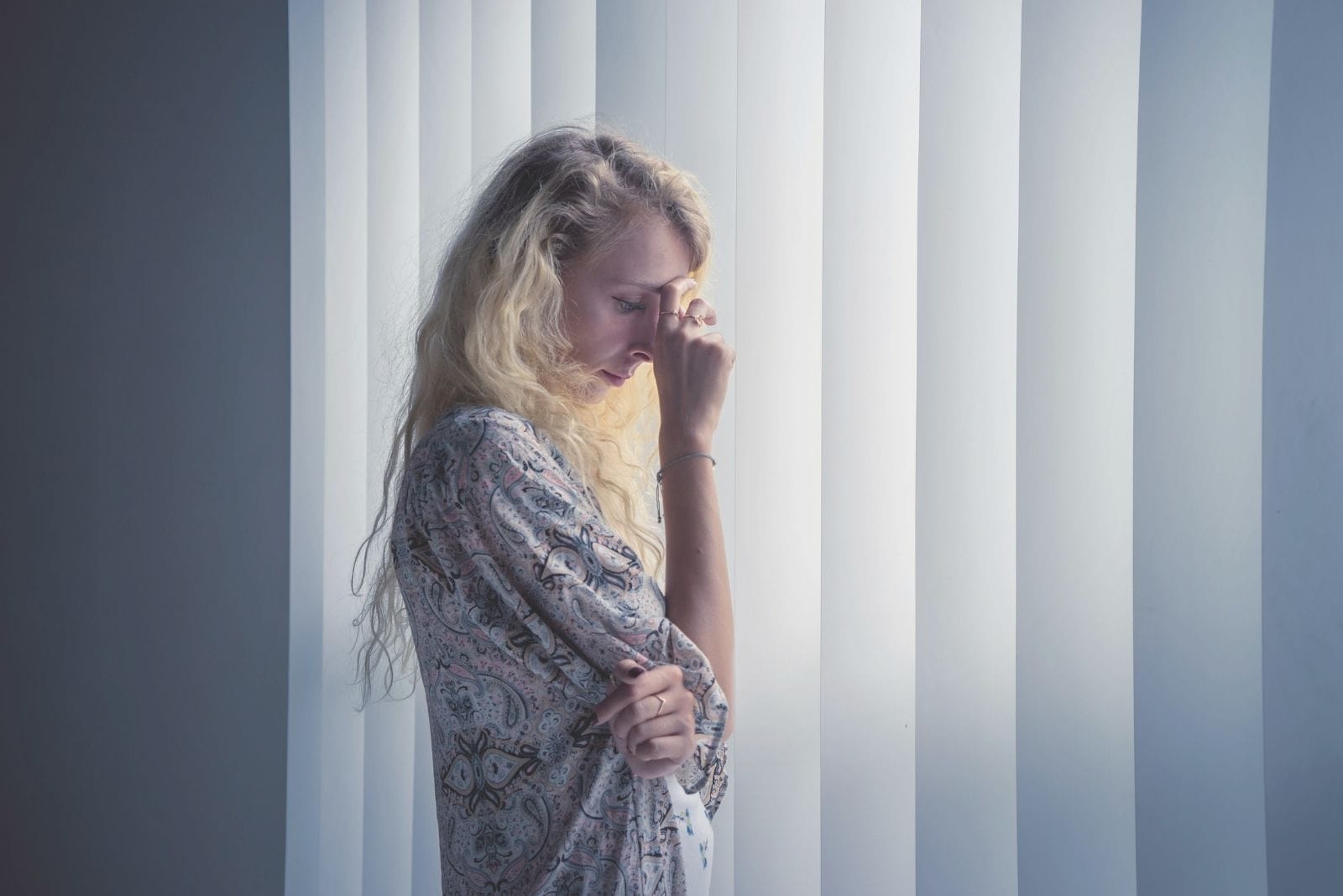 peinzende blonde Kaukasische vrouw die haar hand in haar voorhoofd plaatst en bij de gesloten vensters staat