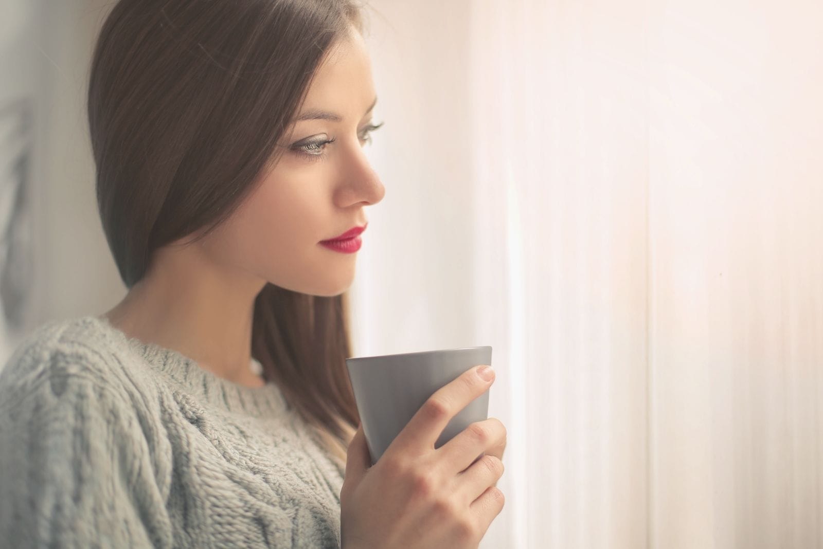 mujer pensativa con una taza en la mano sentada cerca de las ventanas