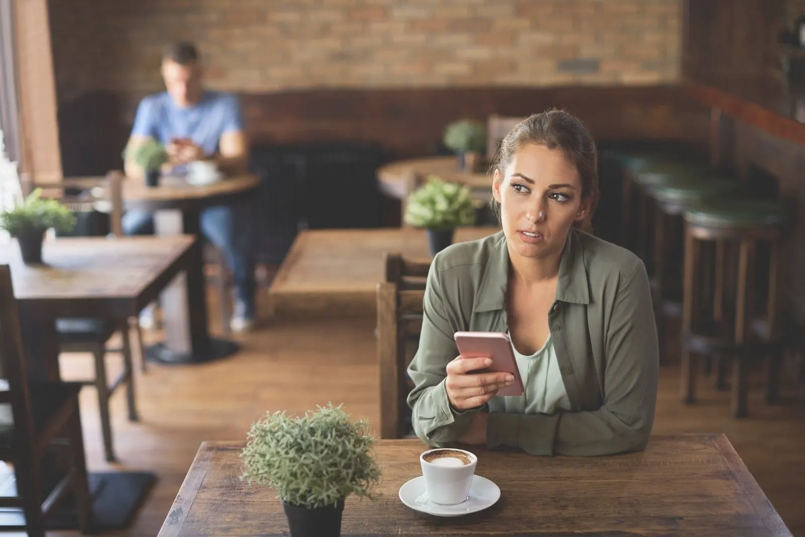 pensive woman text messaging insde cafe with a blurred guy having coffee at the back