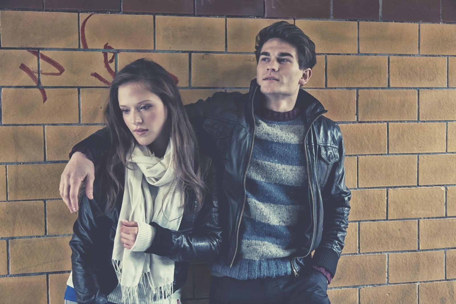 pensive young couple wearing leather jacket standing againg brick wall with hand of the man around the woman's shoulders