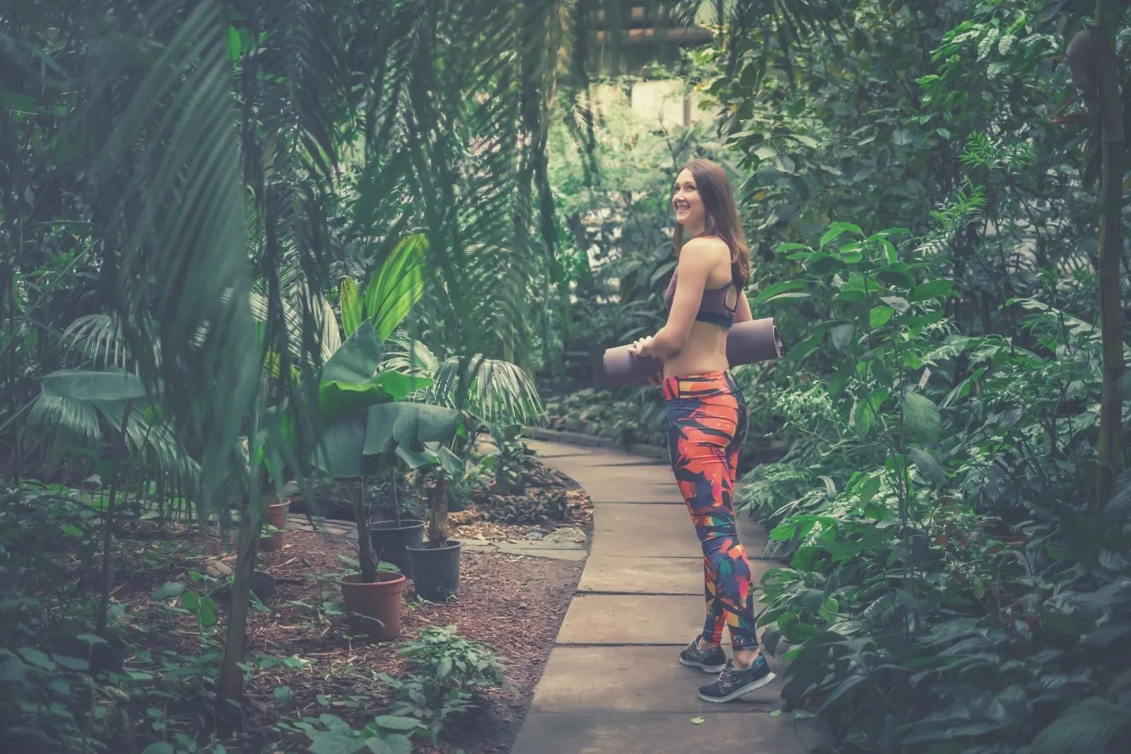 positive female yoga instructor posing at a greenhouse bringing a mat
