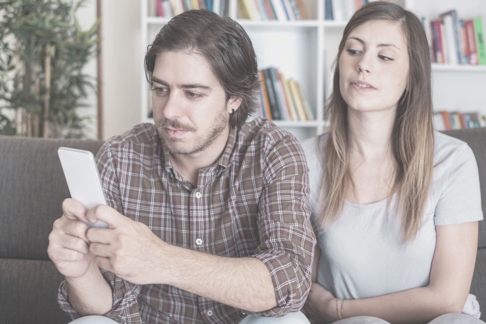 suspicious woman snooping on the man's cellphone while he is texting and sitting beside her in the sofa