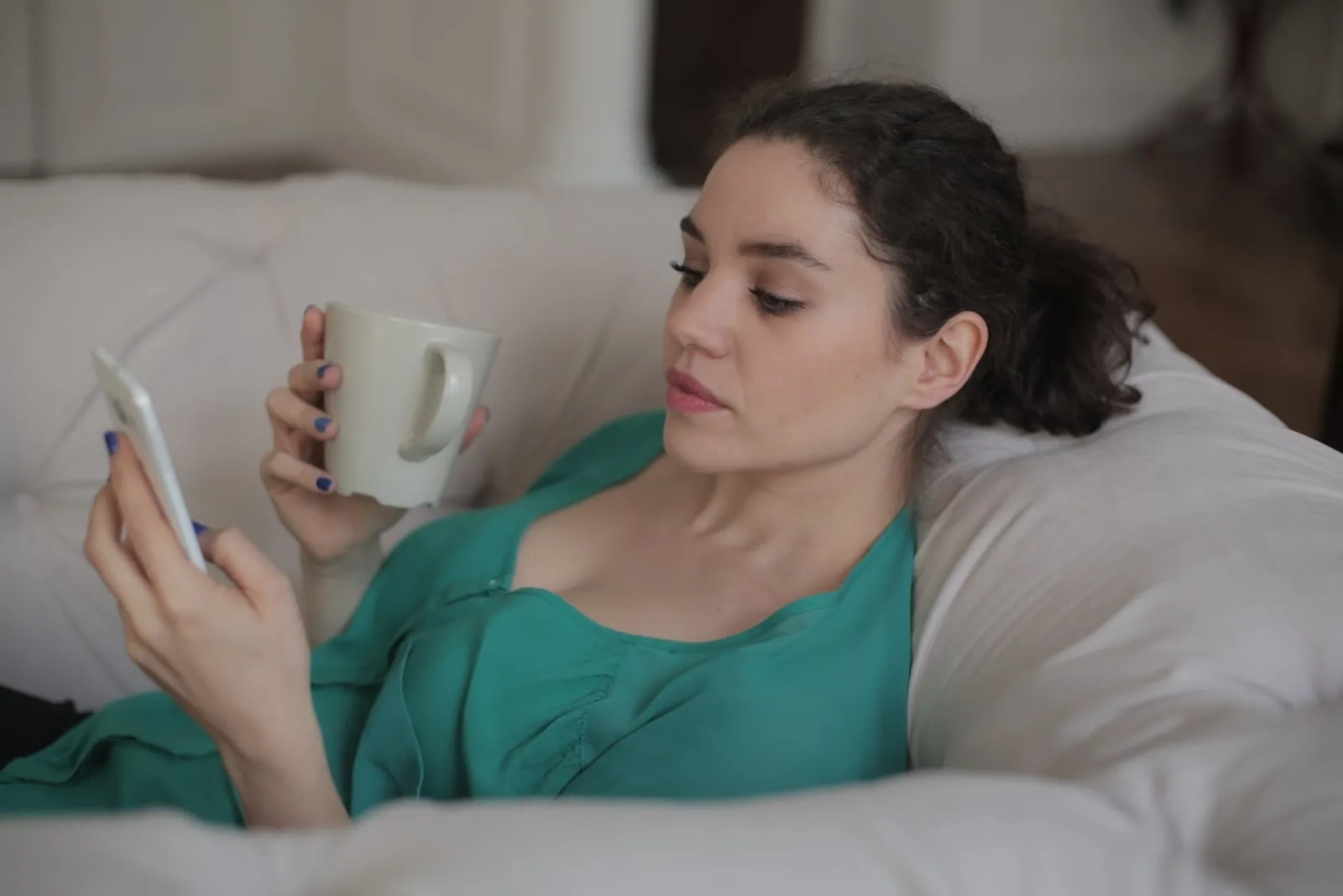 woman with mug looking at phone while sitting on sofa