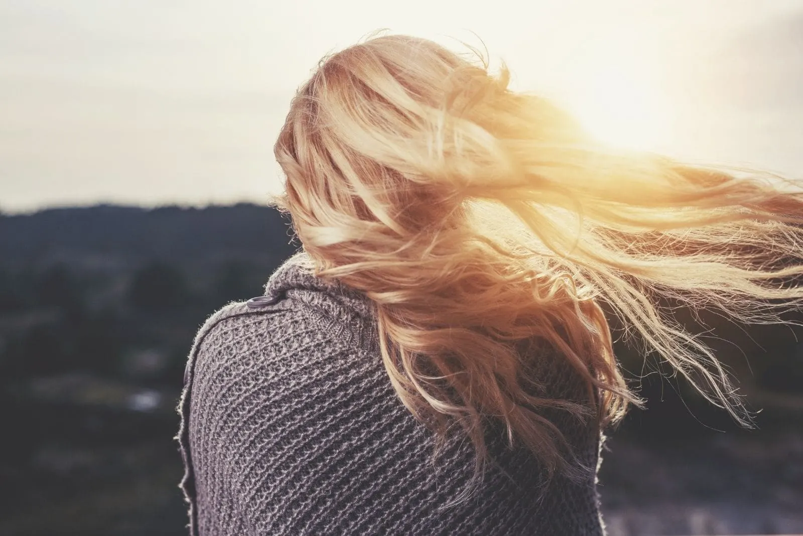woman showing her back while facing the sunrise with blonde hair blown by the wind