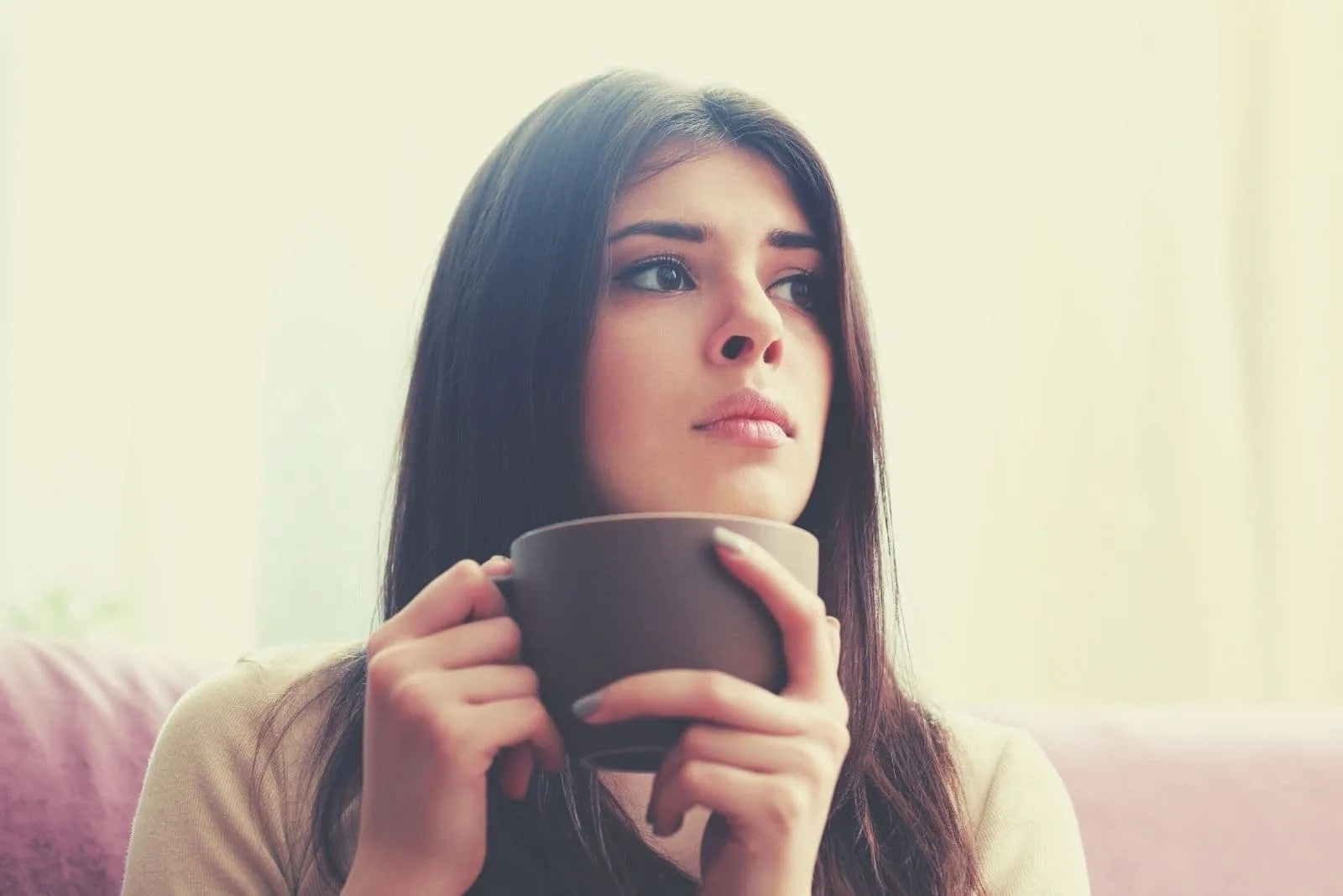young brunette woman thinking deeply while drinking coffee inside home