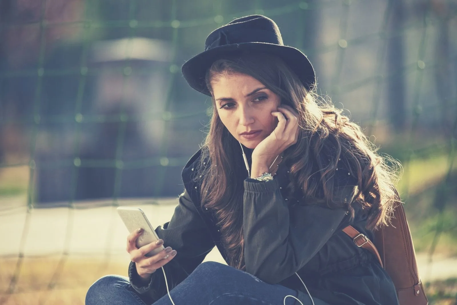 young sad woman with a phone sitting outdoors with a hat
