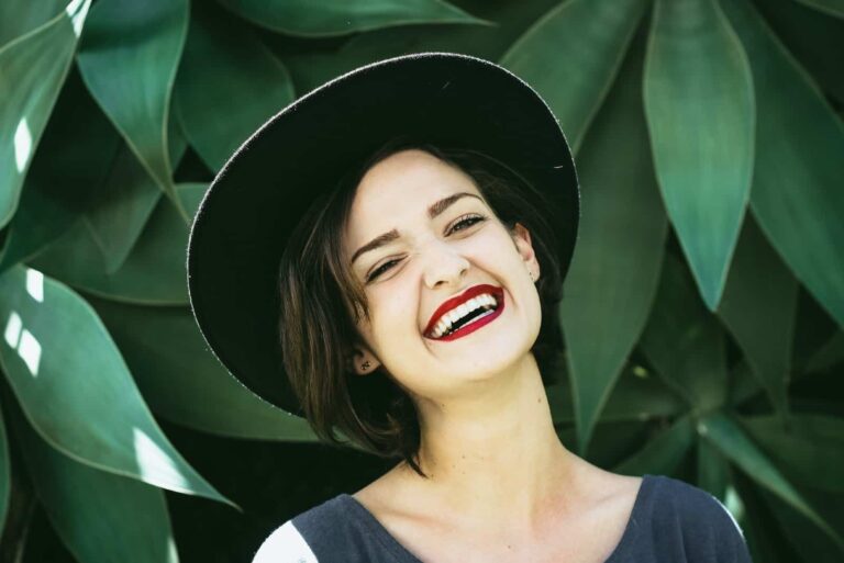 woman with hat smiling while standing near plant